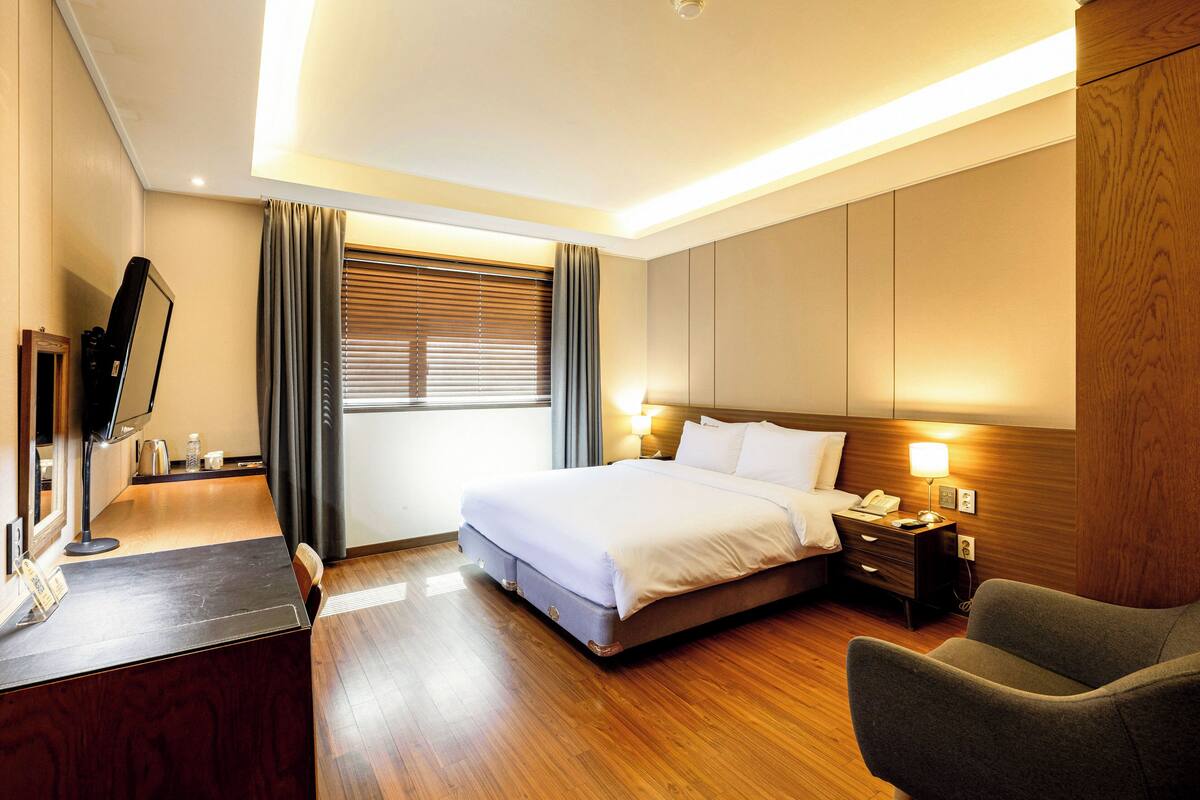 A hotel room with a bed, bedside table, lamp, and a desk with a computer.