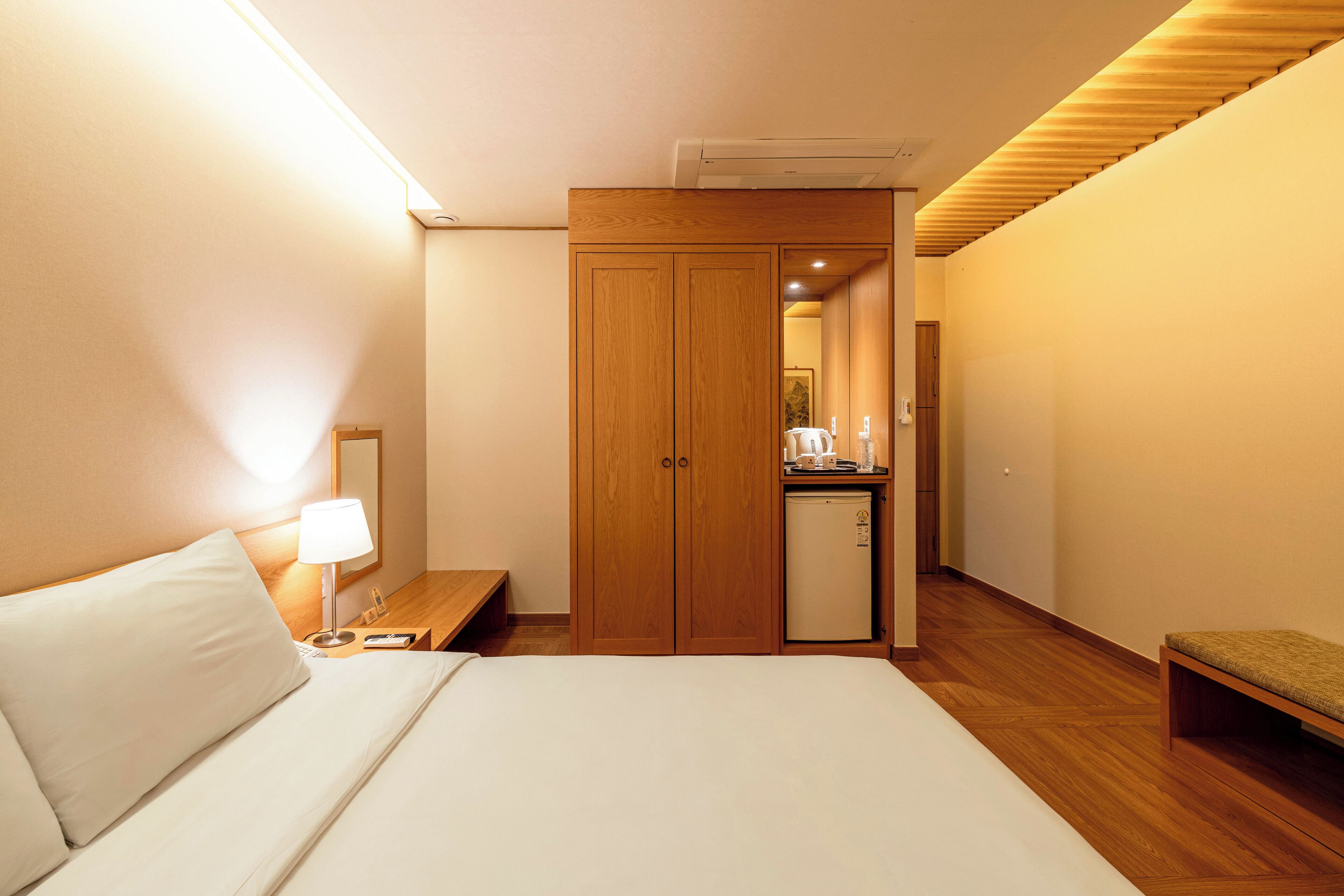 A hotel room with a bed, a desk with a lamp, a wooden wardrobe, and a small kitchenette.