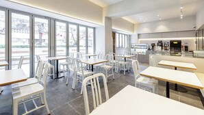 En modern restaurang med vita stolar och bord, stora fönster och ett köksområde som är synligt för gästerna.