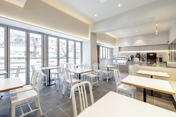 A modern restaurant with white chairs and tables, large windows, and a visible kitchen area.