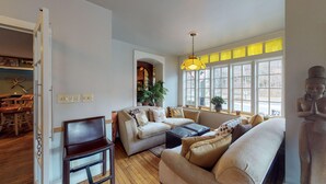 A living room with a sofa, a chair, a table, and a statue. There is a window with yellow curtains and a plant on the windowsill.
