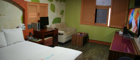 A hotel room with a bed, a desk with a computer, a chair, a small table, a television, and a window with blinds.