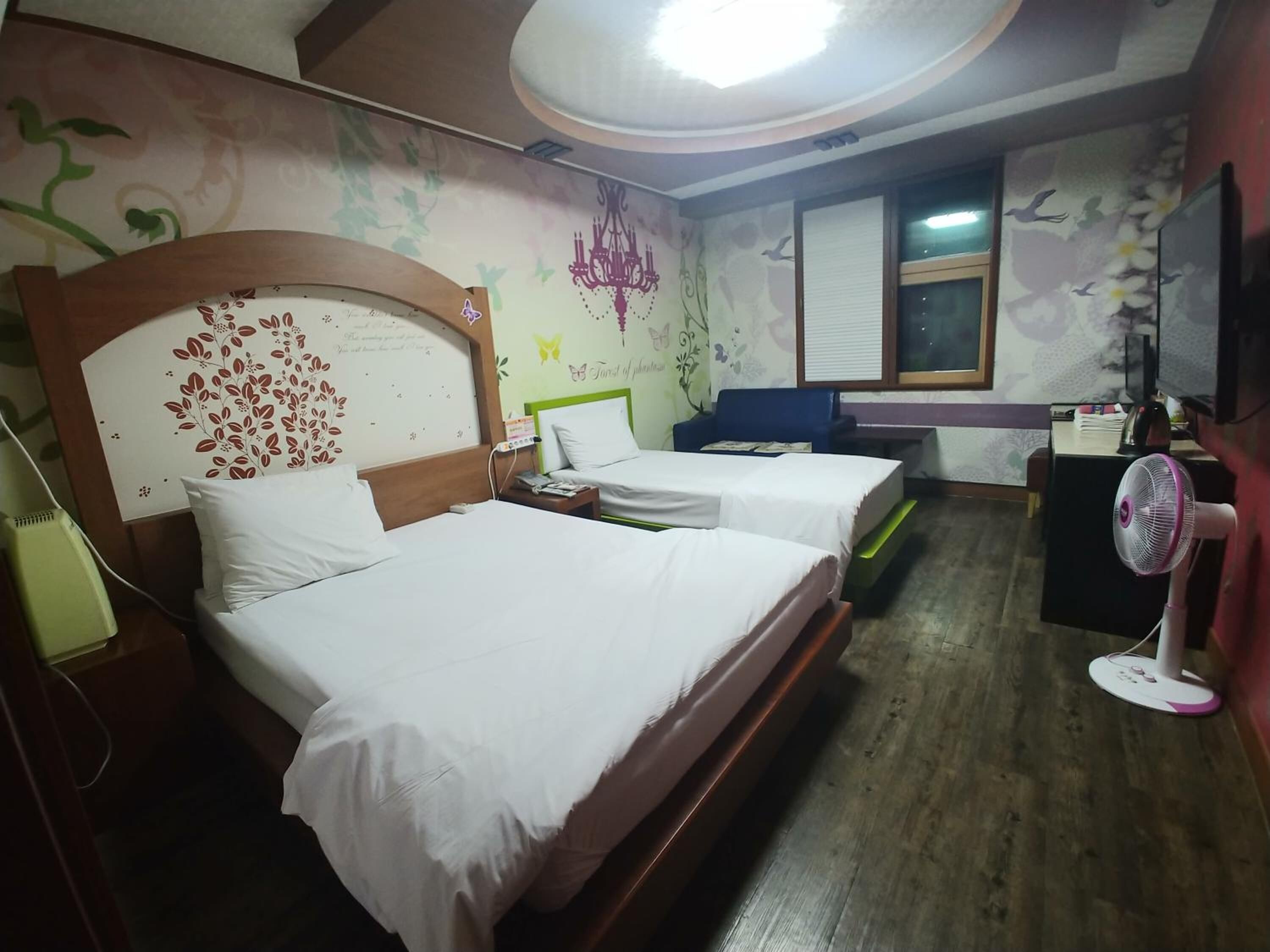 A hotel room with two beds, a desk, a chair, a standing fan, and a window with blinds.