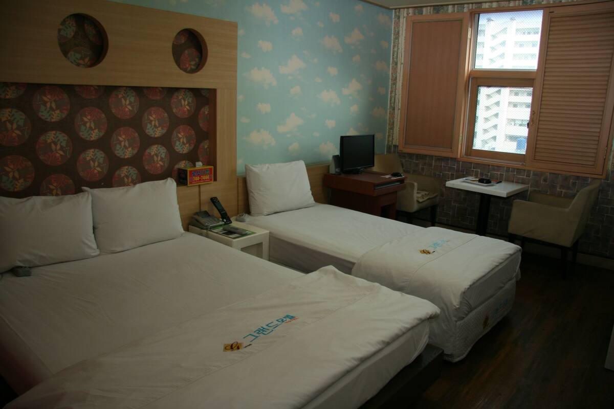 Una habitación de hotel con dos camas, un escritorio con ordenador, televisor y ventana con persianas.