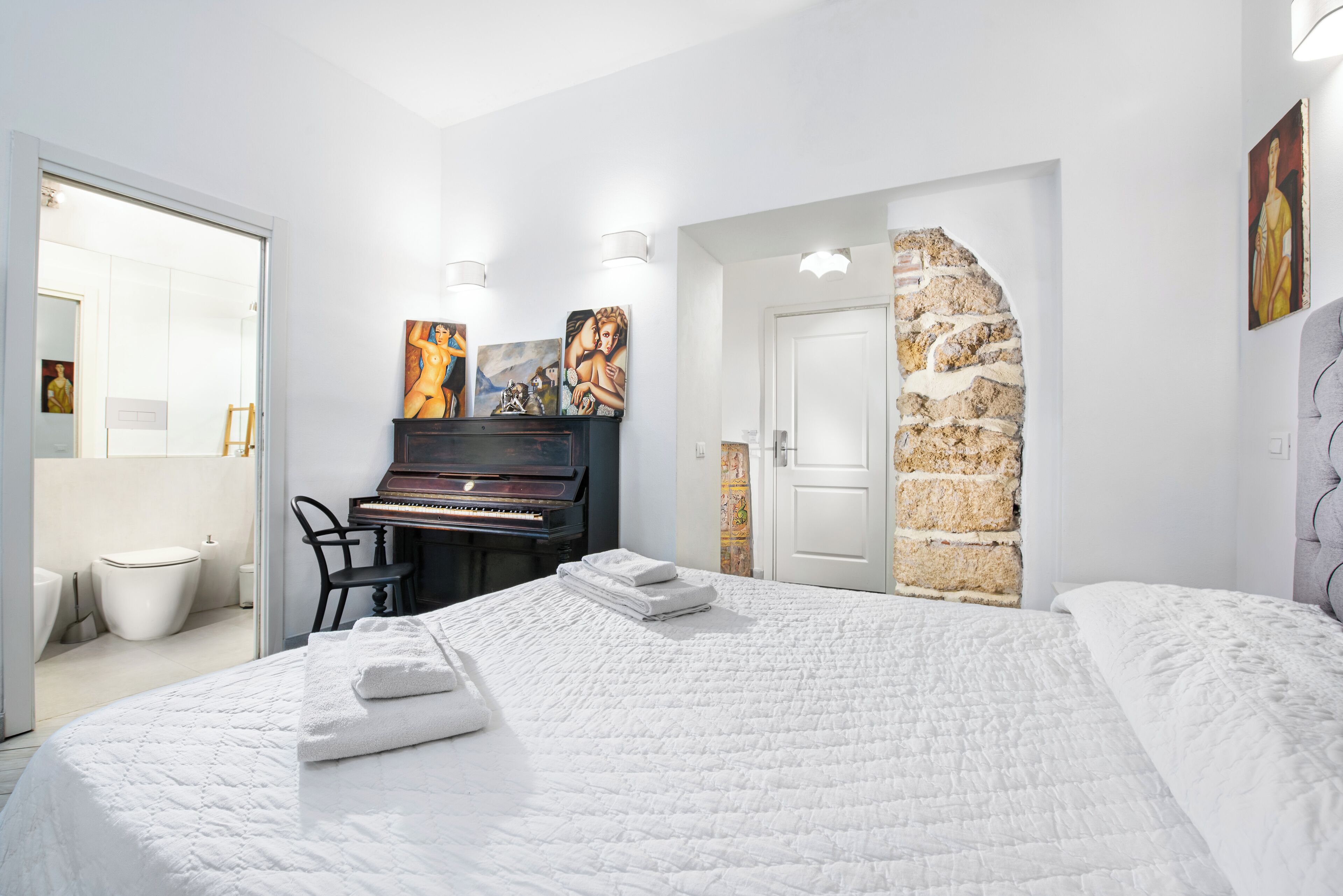 Une chambre avec un piano, un lit recouvert de linge de lit blanc et des œuvres d’art accrochées aux murs.
