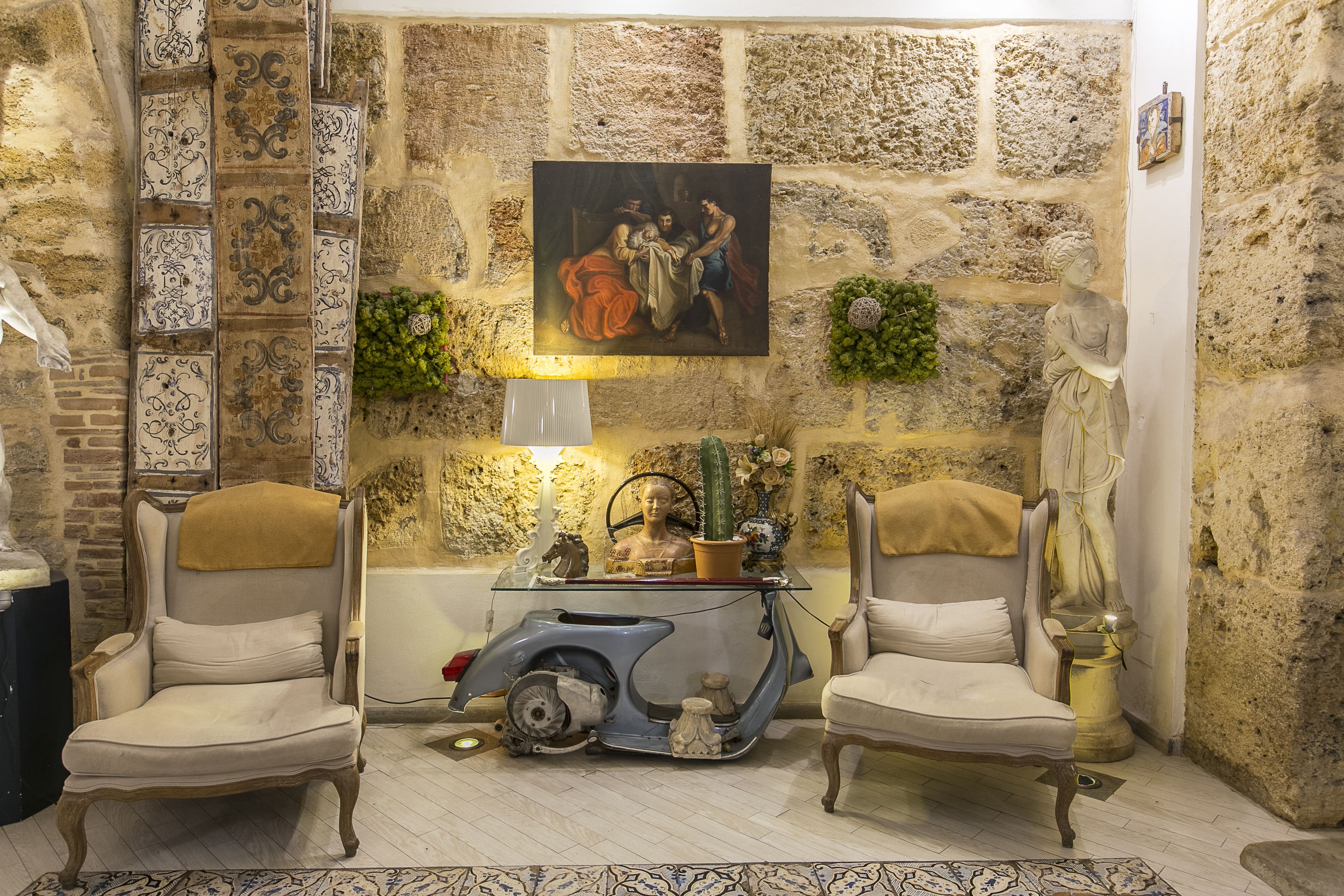 A room with stone walls, a painting, two armchairs, a vintage scooter, and a small table with decorative items.