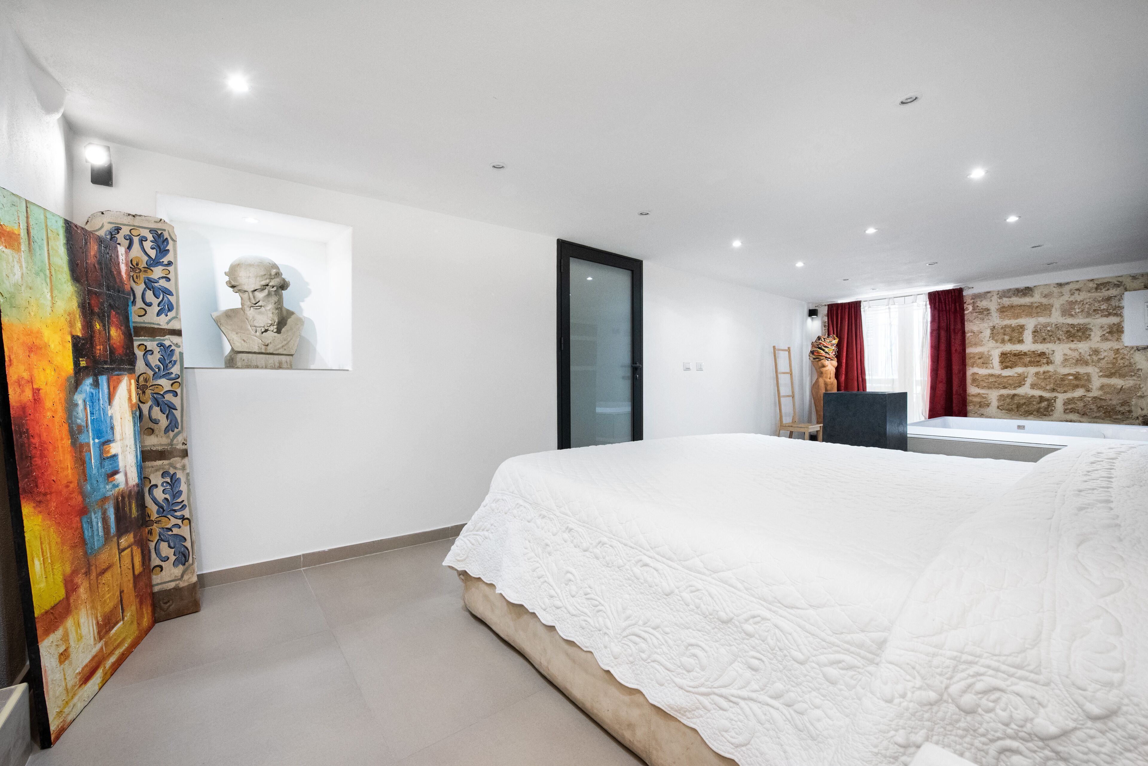 Un dormitorio con una cama grande, una pared de piedra, una ventana con cortinas rojas y un cuadro a la izquierda.