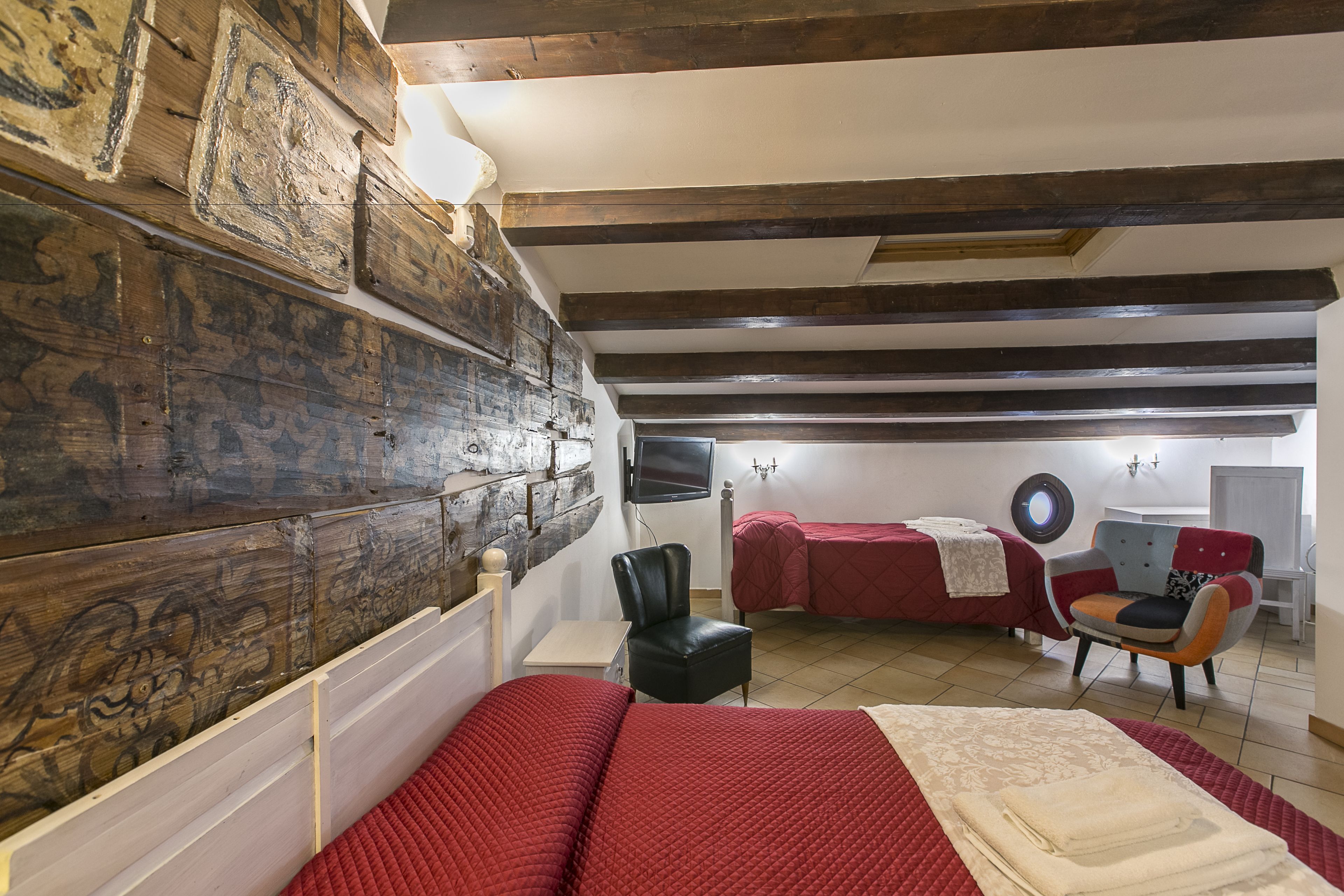 A room with a wooden wall, a bed with a red bedspread, a chair, and a small table.