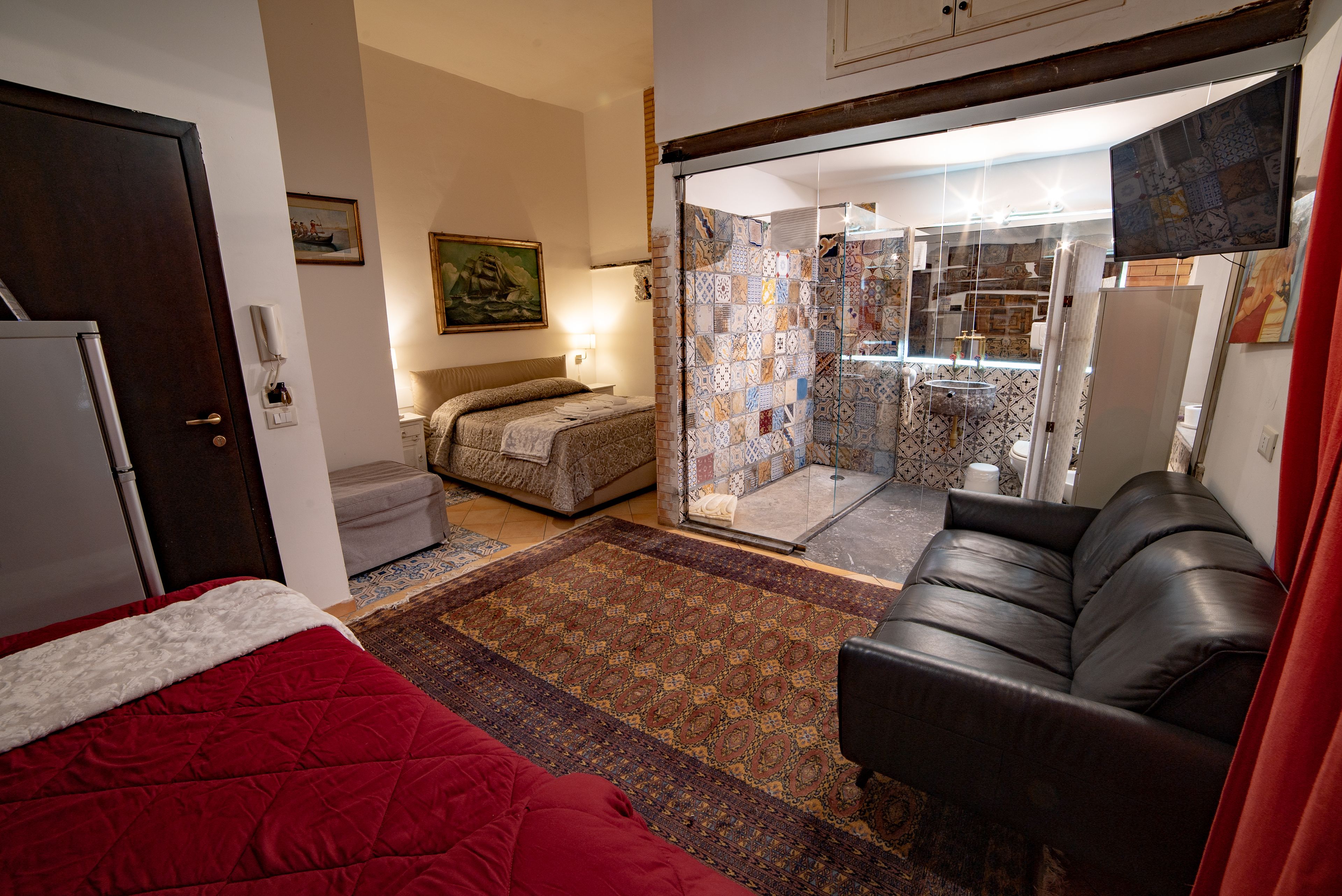 Une chambre avec un lit rouge, un canapé en cuir noir, une petite table et une porte en verre donnant sur une salle de bain.