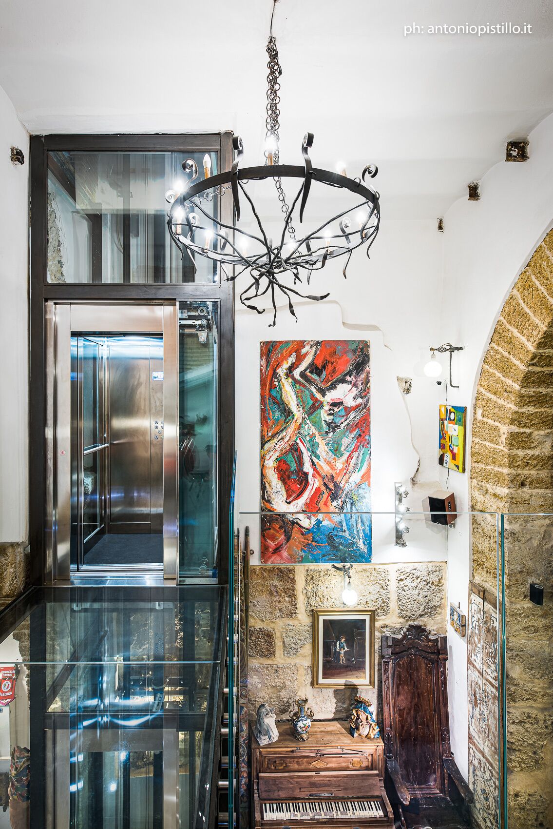 Un intérieur moderne avec un ascenseur en verre, un lustre, des œuvres d’art abstrait et un piano.