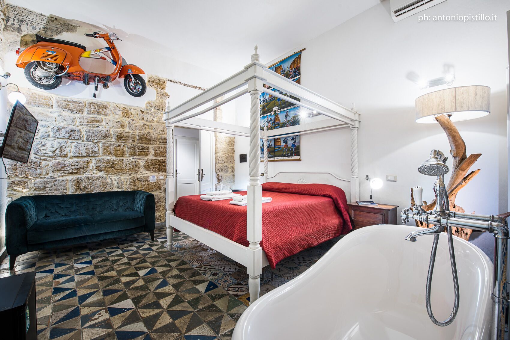 Ein Bett mit Baldachin und roter Bettdecke, ein orangefarbener, alter Roller, eine Steinmauer, ein blaues Sofa und eine Badewanne mit Duschkopf.