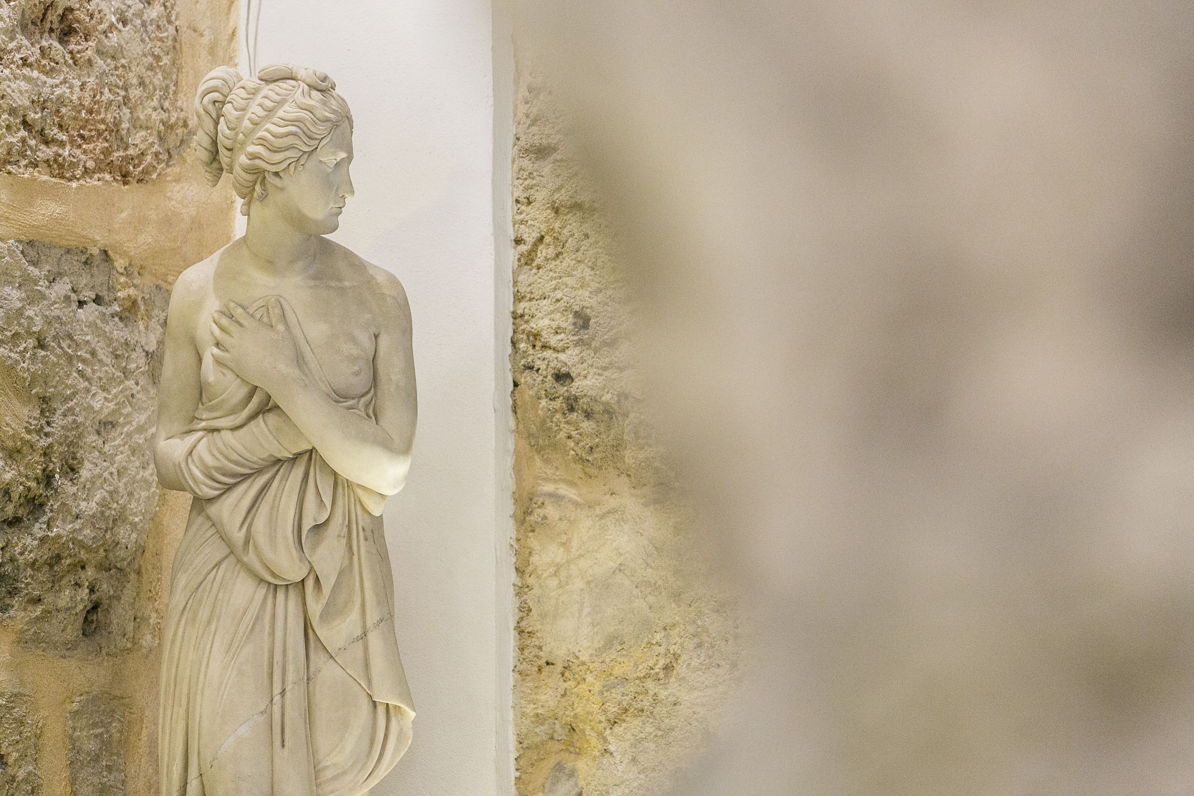 A marble statue of a woman with a draped garment, positioned against a textured wall.