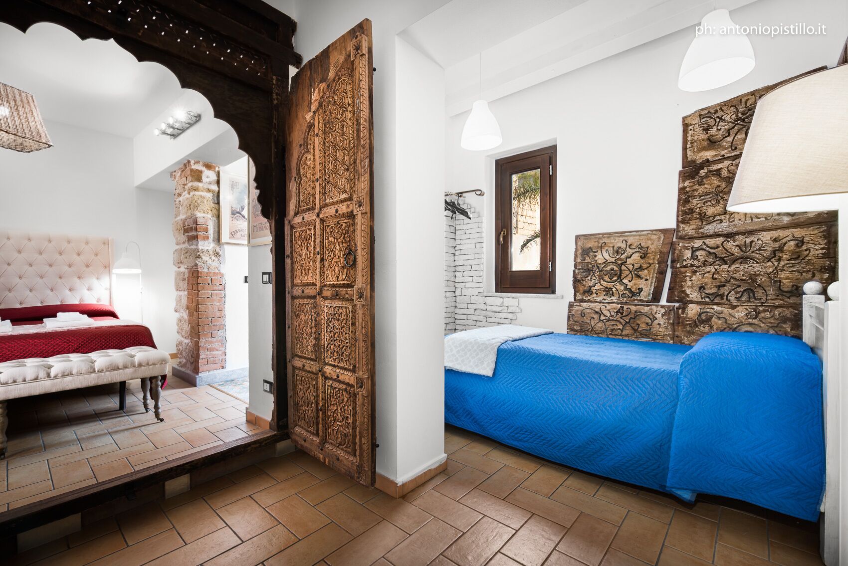 A bedroom with a large wooden door, a bed with a red and white headboard, and a blue bedspread.
