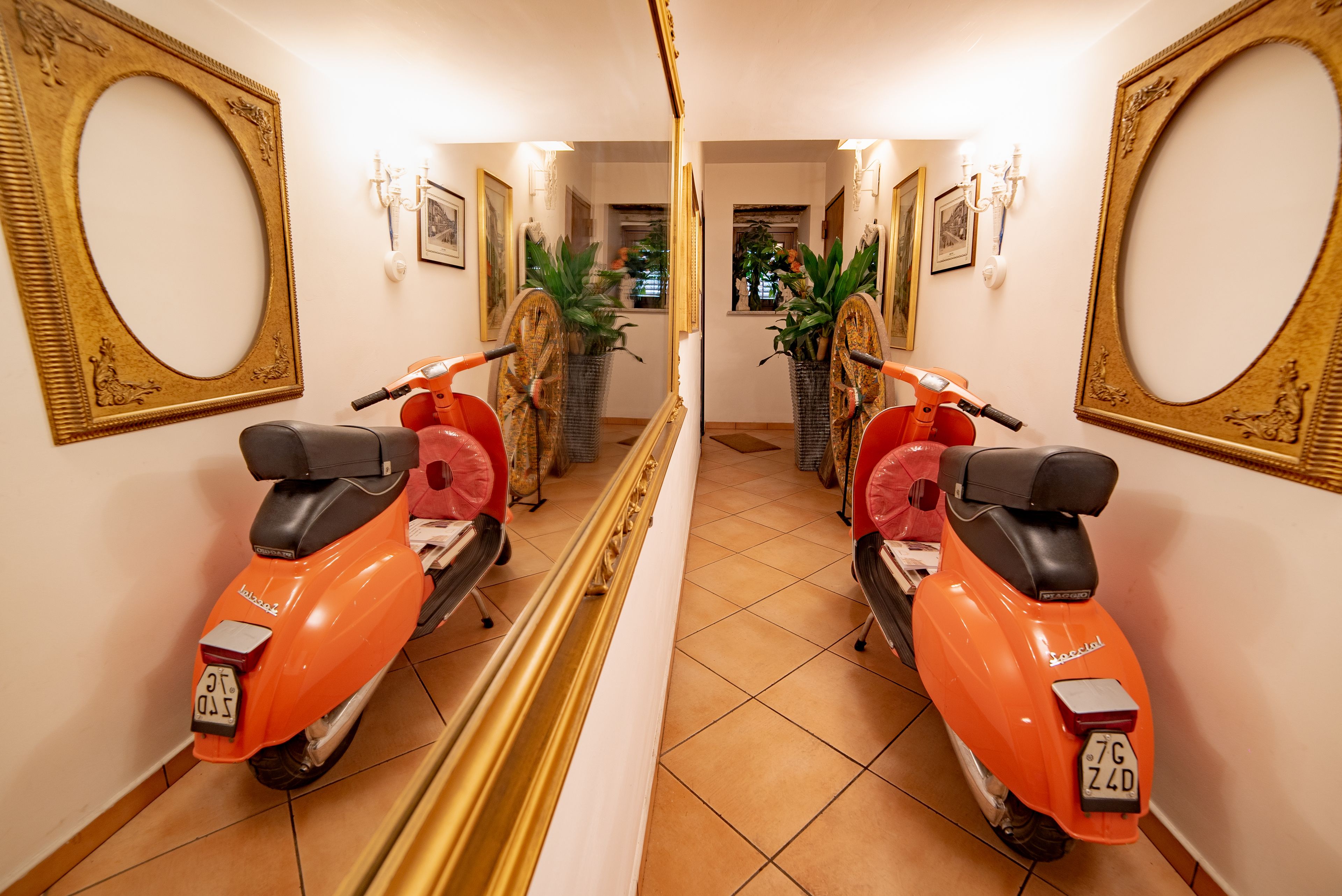 A hallway with two orange scooters parked on either side, a mirror reflecting the room, and a potted plant in the center.