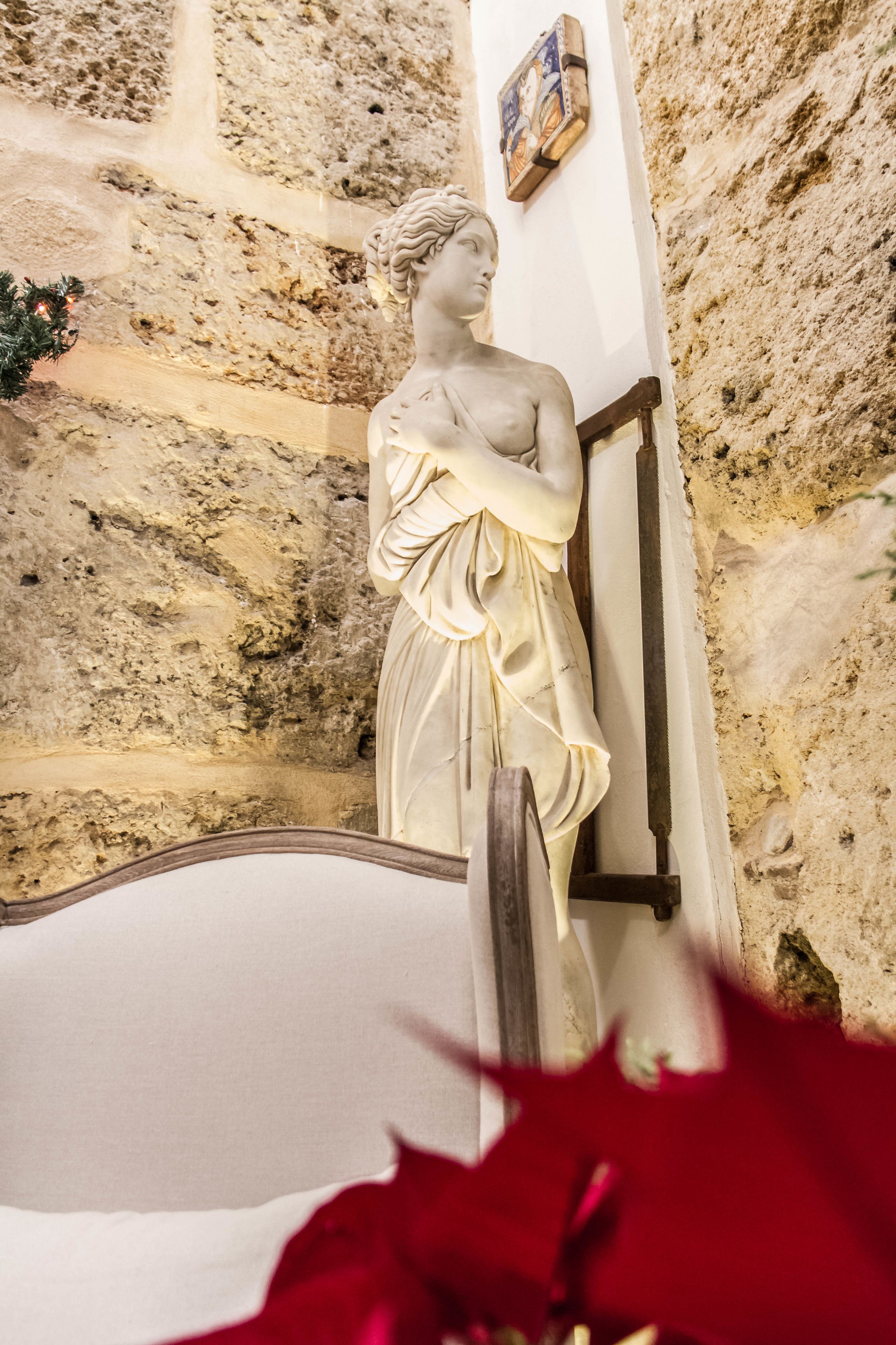 A statue of a woman in a white dress standing next to a wooden chair, with a stone wall and a framed picture in the background.