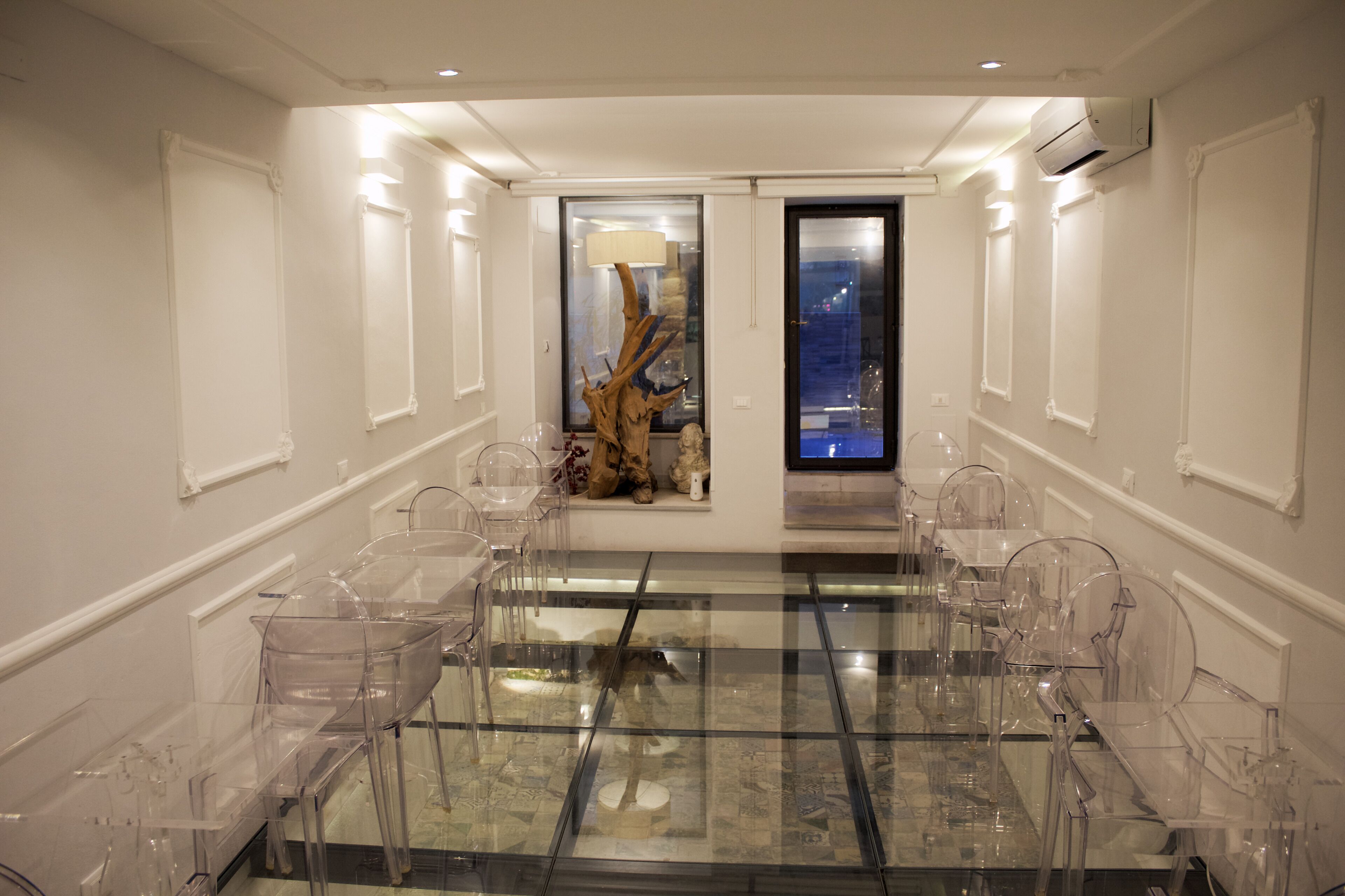 A modern conference room with a glass table and clear chairs, a decorative tree sculpture, and a city view through a glass door.