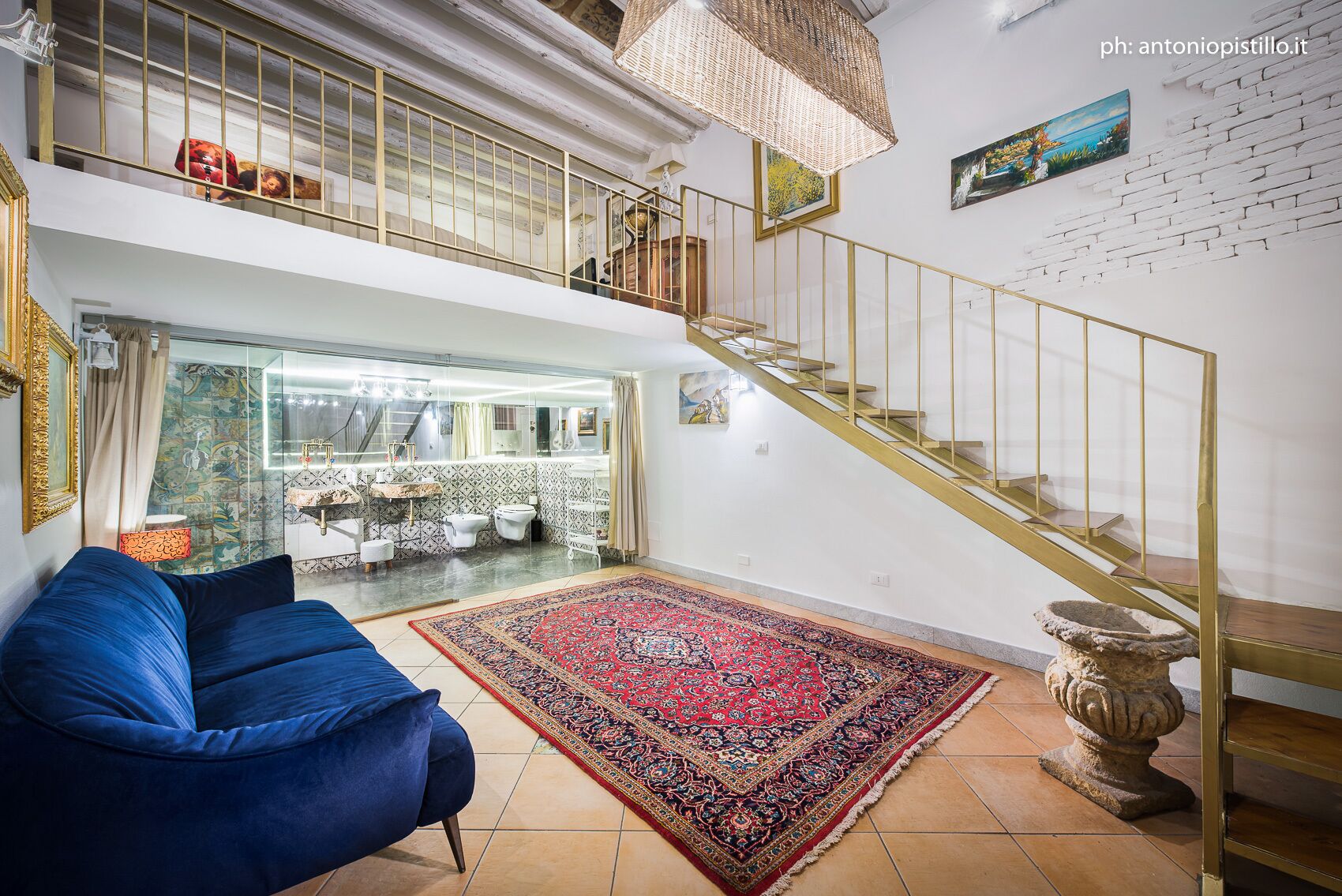 Un salón amplio con un sofá azul, un tapete grande con estampado, una escalera con barandilla dorada y vistas a una cocina y un baño a través de un separador de vidrio.