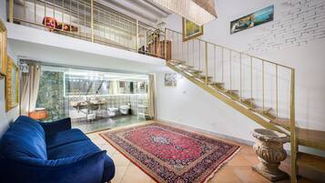 Un salón amplio con un sofá azul, un tapete grande con estampado, una escalera con barandilla dorada y vistas a una cocina y un baño a través de un separador de vidrio.