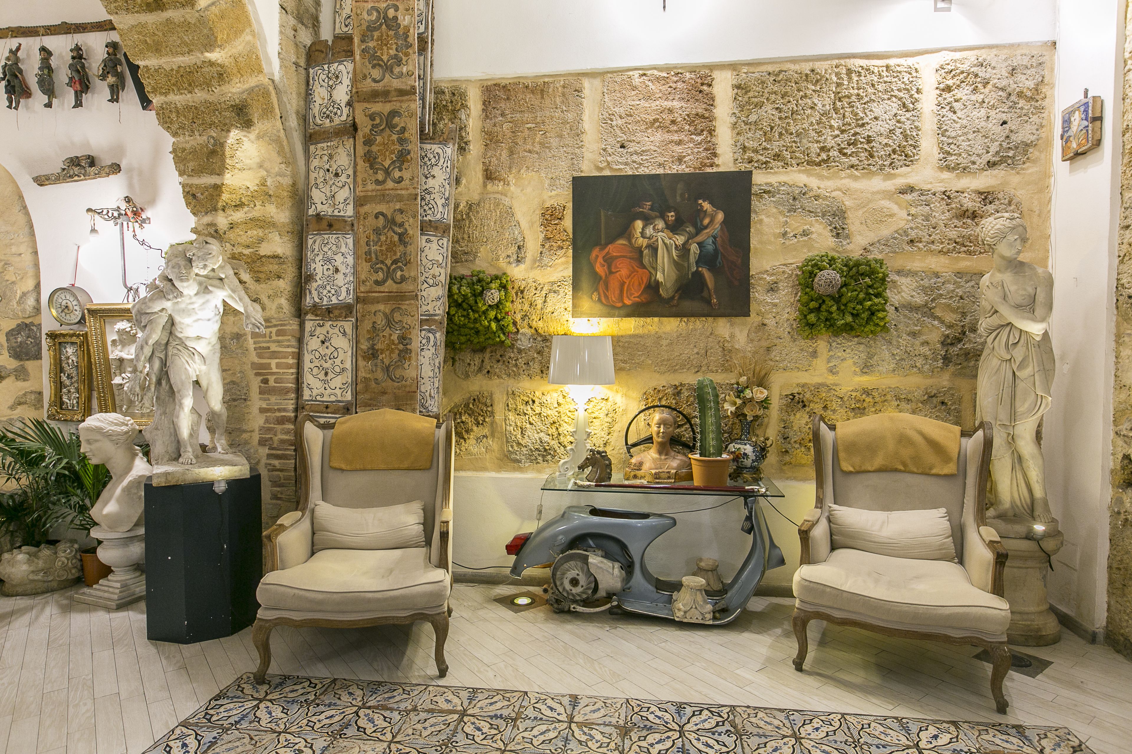 Een kamer met stenen muren, twee fauteuils, een oude scooter en diverse beelden en decoraties.
