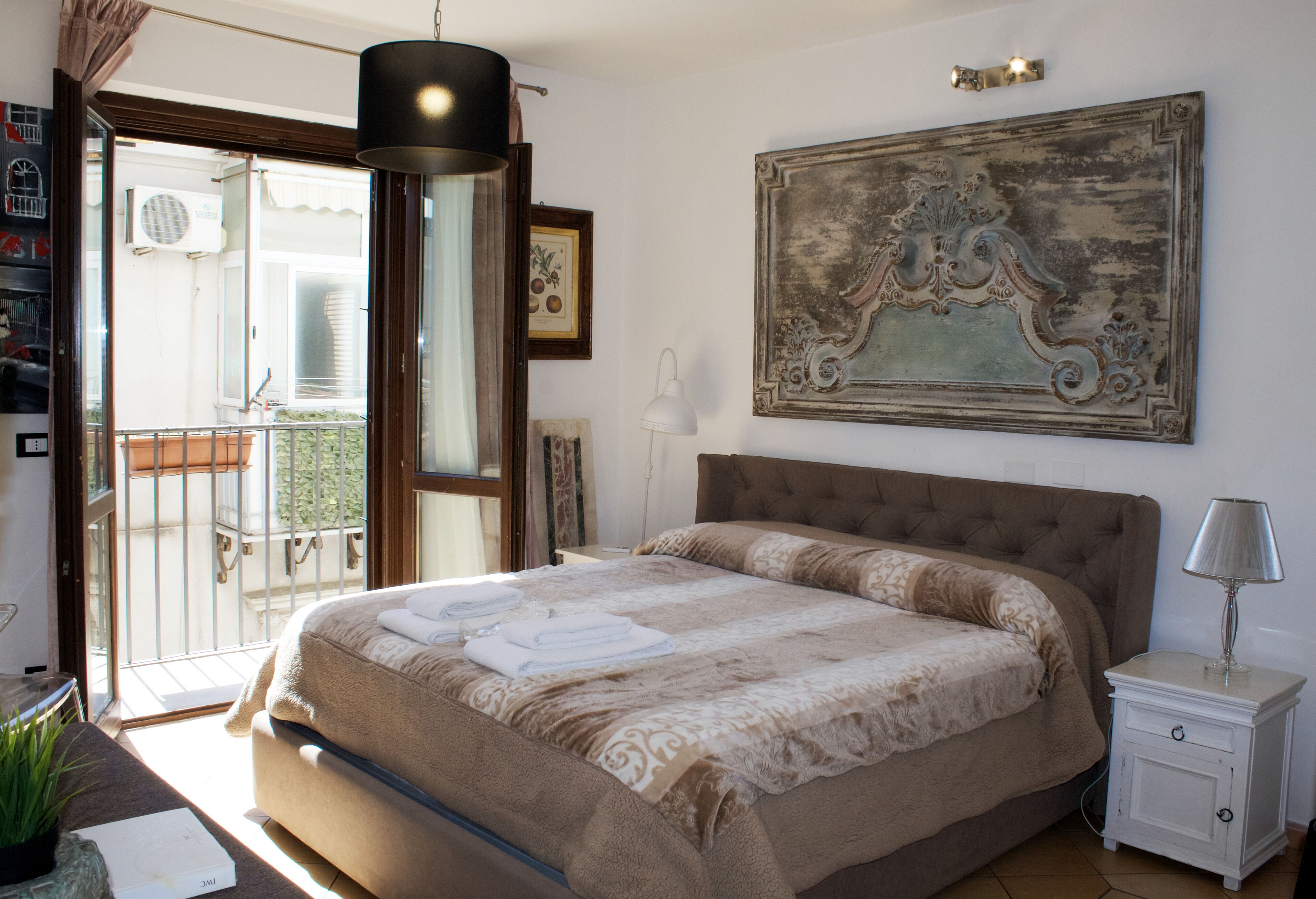 Un dormitorio con una cama grande, una mesita de noche, una lámpara, una ventana con vistas y un cuadro colgado en la pared.