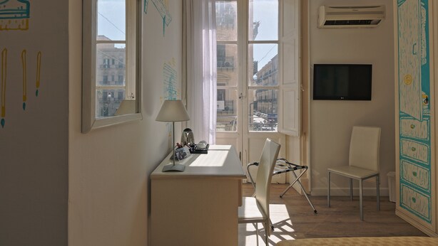 A room with a desk, chair, and a television. There are windows with curtains, an air conditioning unit, and a view of buildings outside. - Afea Art & Rooms (Palermo)