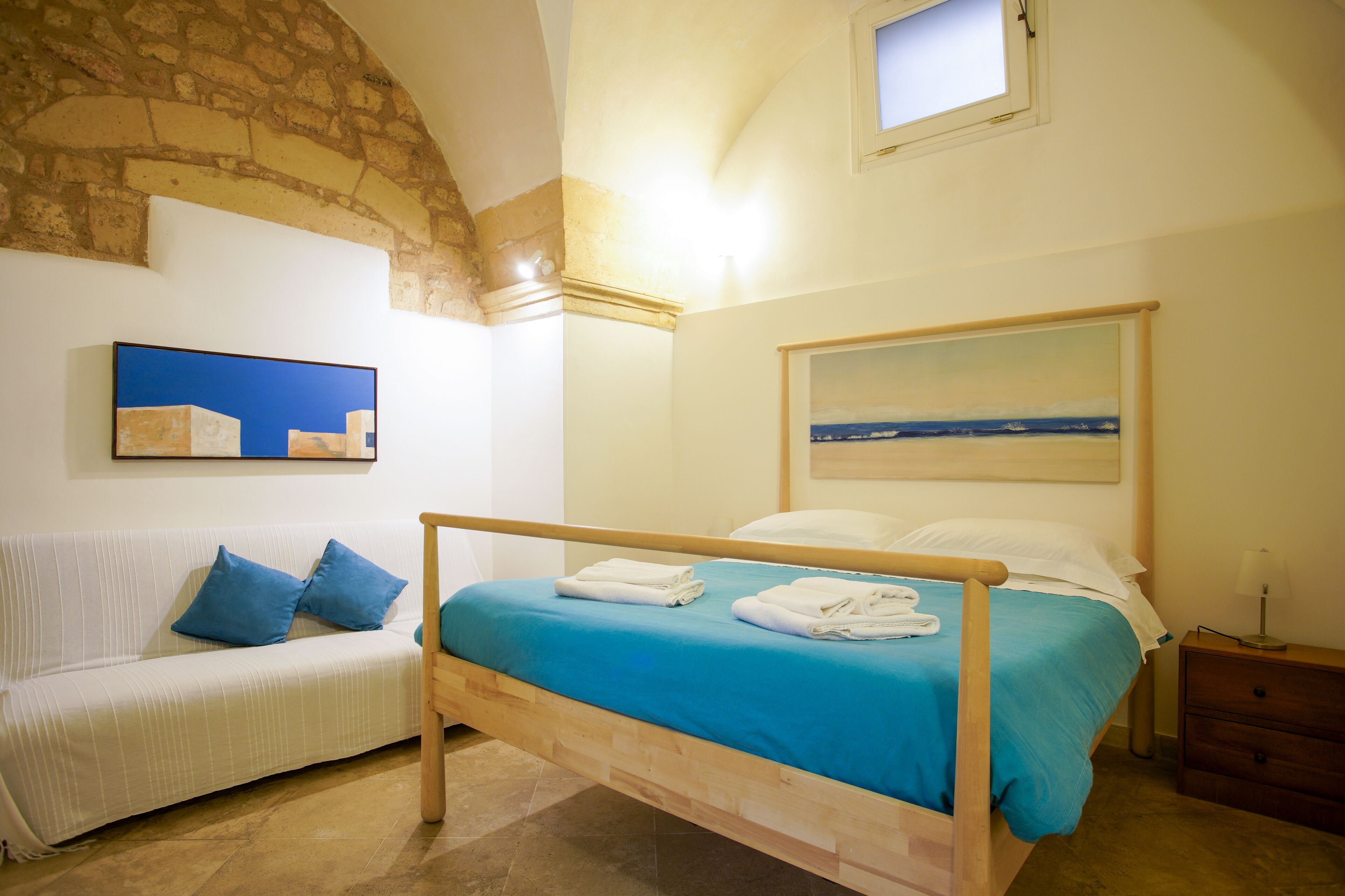 Un dormitorio con cama, mesita de noche, banco de madera, un cuadro y pared de piedra.