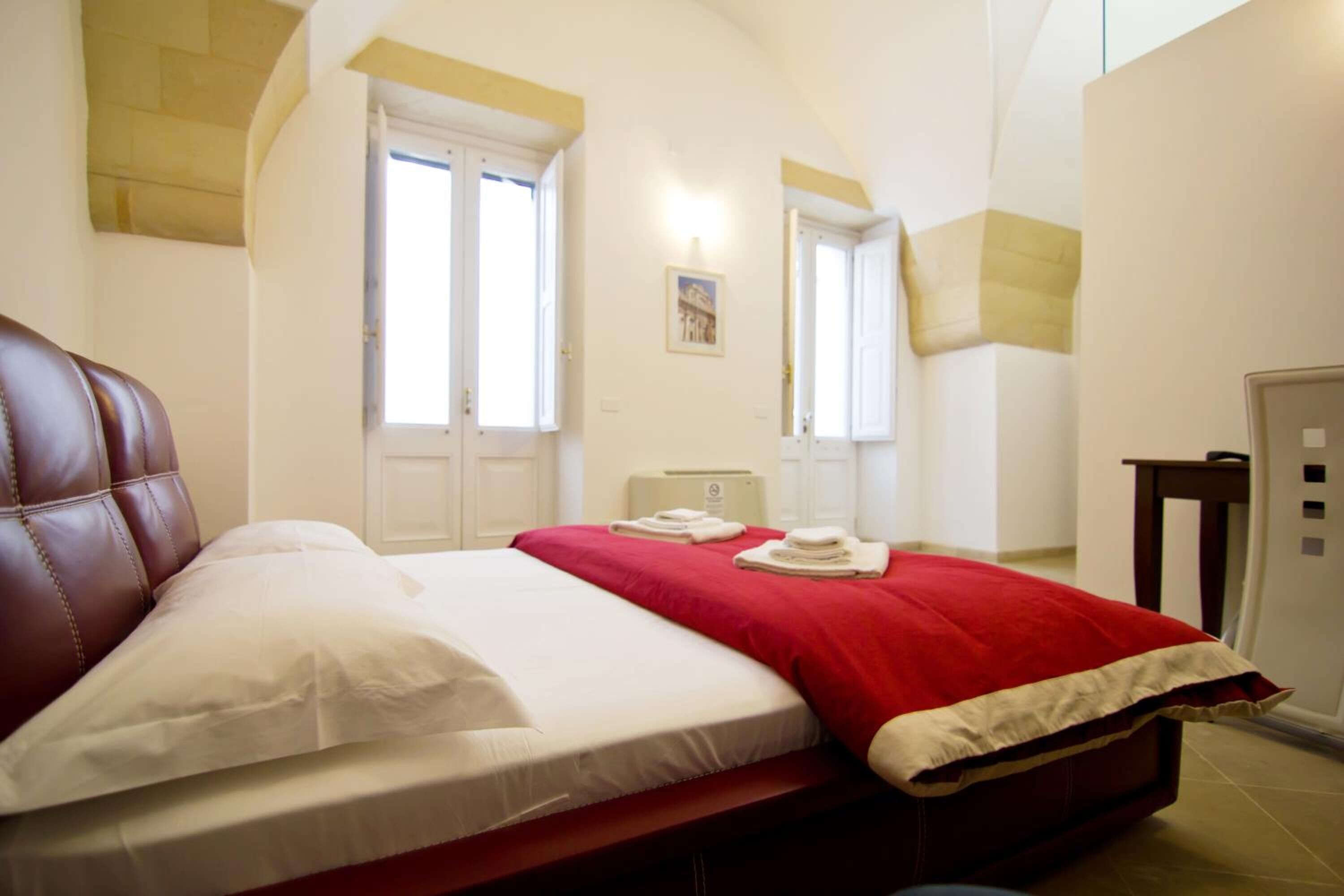 A bedroom with a bed, a red blanket, and a white pillow. There are two doors and a small table with a lamp.