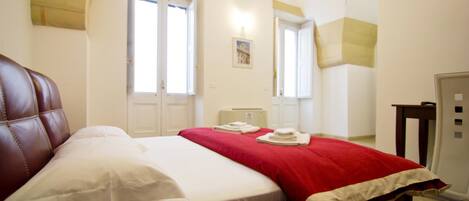 A bedroom with a bed, a red blanket, and a white pillow. There are two doors and a small table with a lamp.