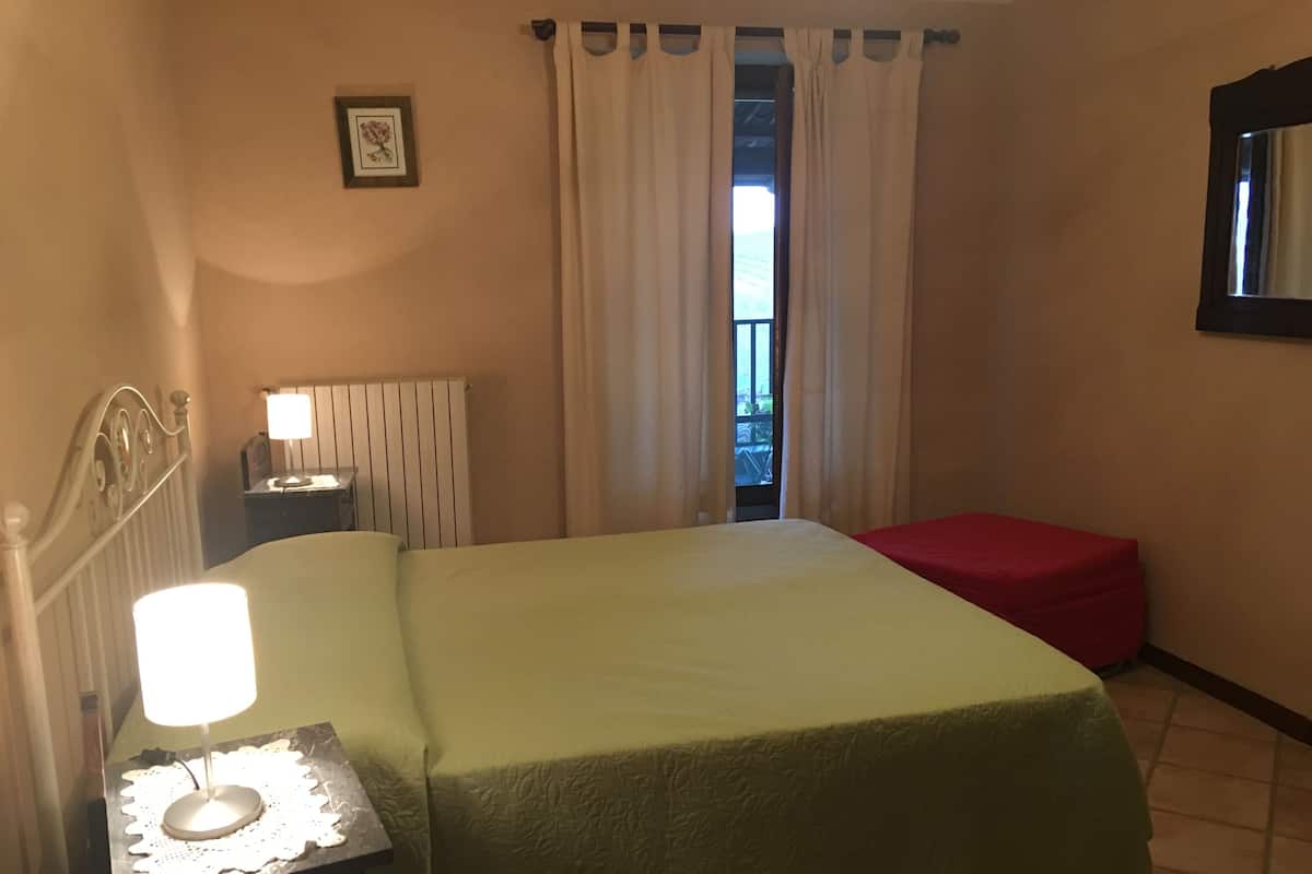 Une chambre d’hôtel avec un lit, une table de chevet, une lampe, un banc rouge, un tableau encadré au mur et une fenêtre avec des rideaux.