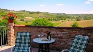 A balcony with two checkered lounge chairs, a small round table with a bottle and glasses, and a view of rolling hills. - Agriturismo La Mussia (Castelnuovo Calcea)