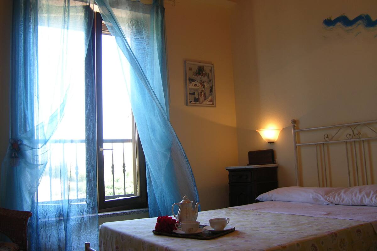 Une chambre à coucher avec un lit, une table de chevet, une lampe, une fenêtre avec des rideaux et un mur orné d’un tableau.