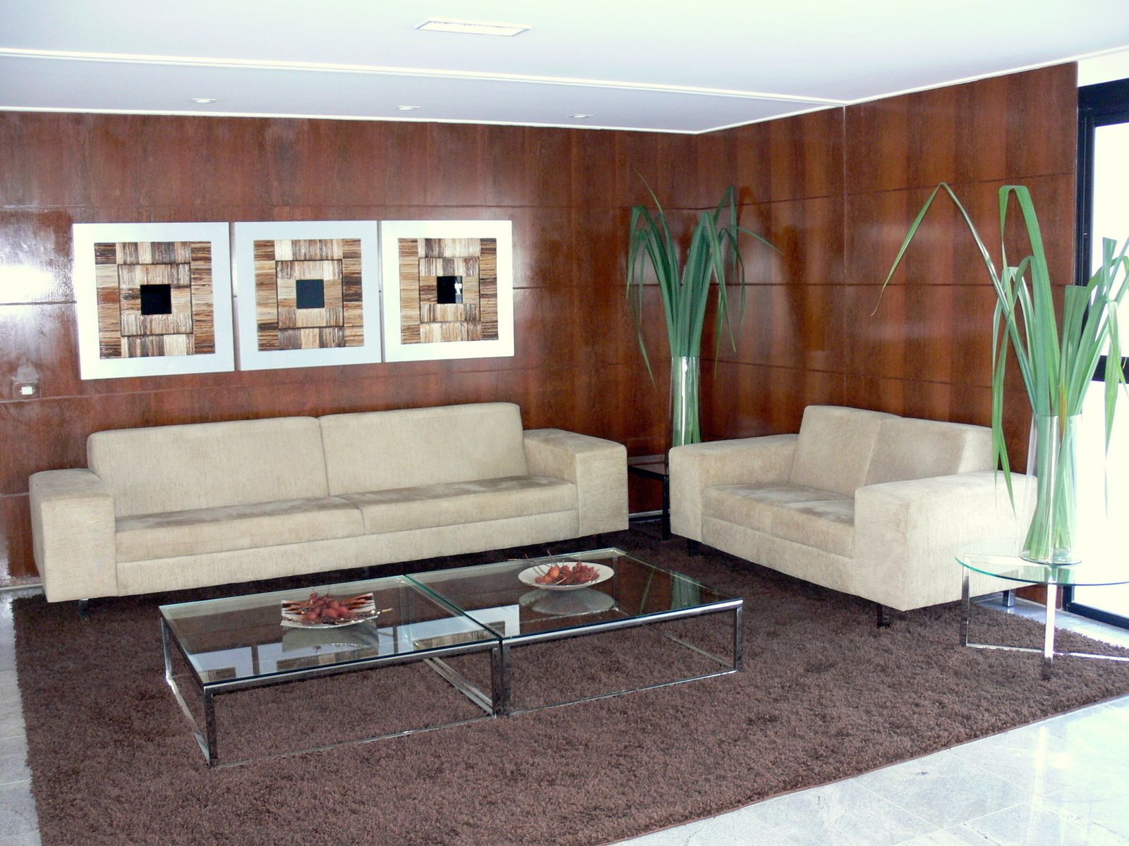 Una sala de estar moderna con un sofá beige, una mesa de centro de vidrio y dos mesas auxiliares. Hay tres cuadros enmarcados en la pared y dos plantas verdes altas.