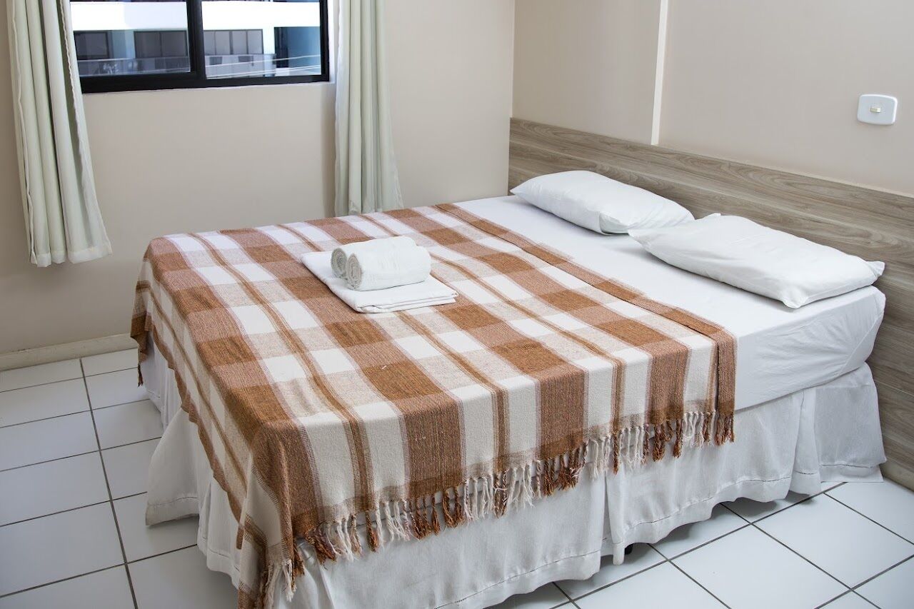 A neatly made bed with a checkered bedspread and a rolled towel, a window with curtains, and a light switch on the wall.