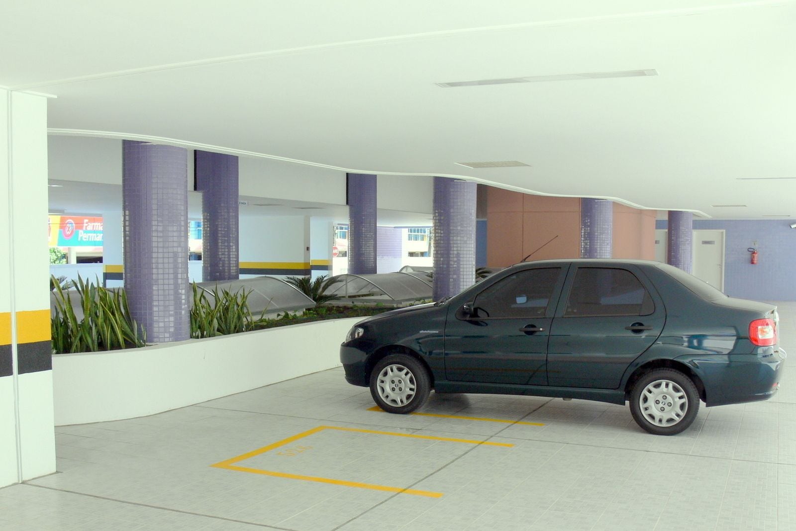 Un estacionamiento con un coche, paredes blancas, pilares morados y un letrero que dice 'Farmacia Perma'.