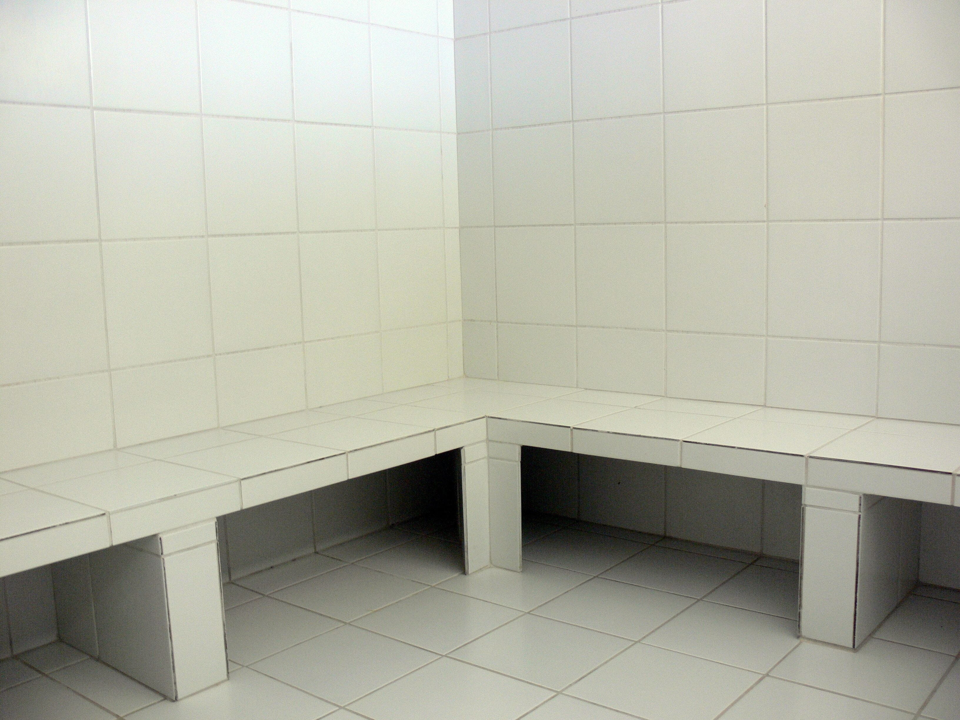 Ein Eckbereich eines Badezimmers mit weiß gefliesten Wänden und Boden, einer Sitzbank und einem Waschbecken.