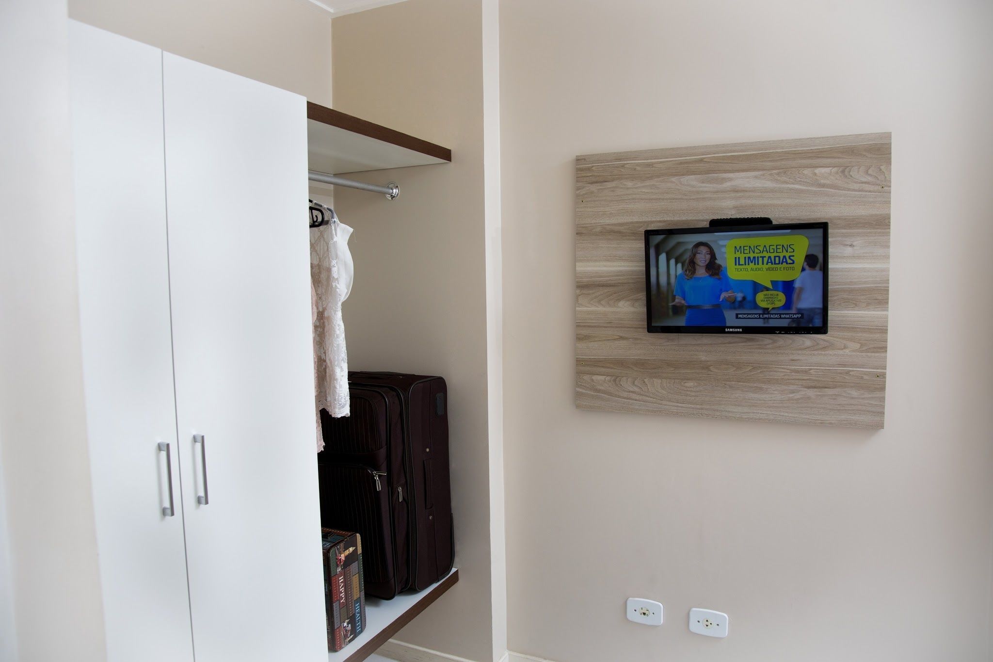 Ein Zimmer mit Kleiderschrank, Bücherregal und an der Wand montiertem Fernseher, der ein Programm zeigt.