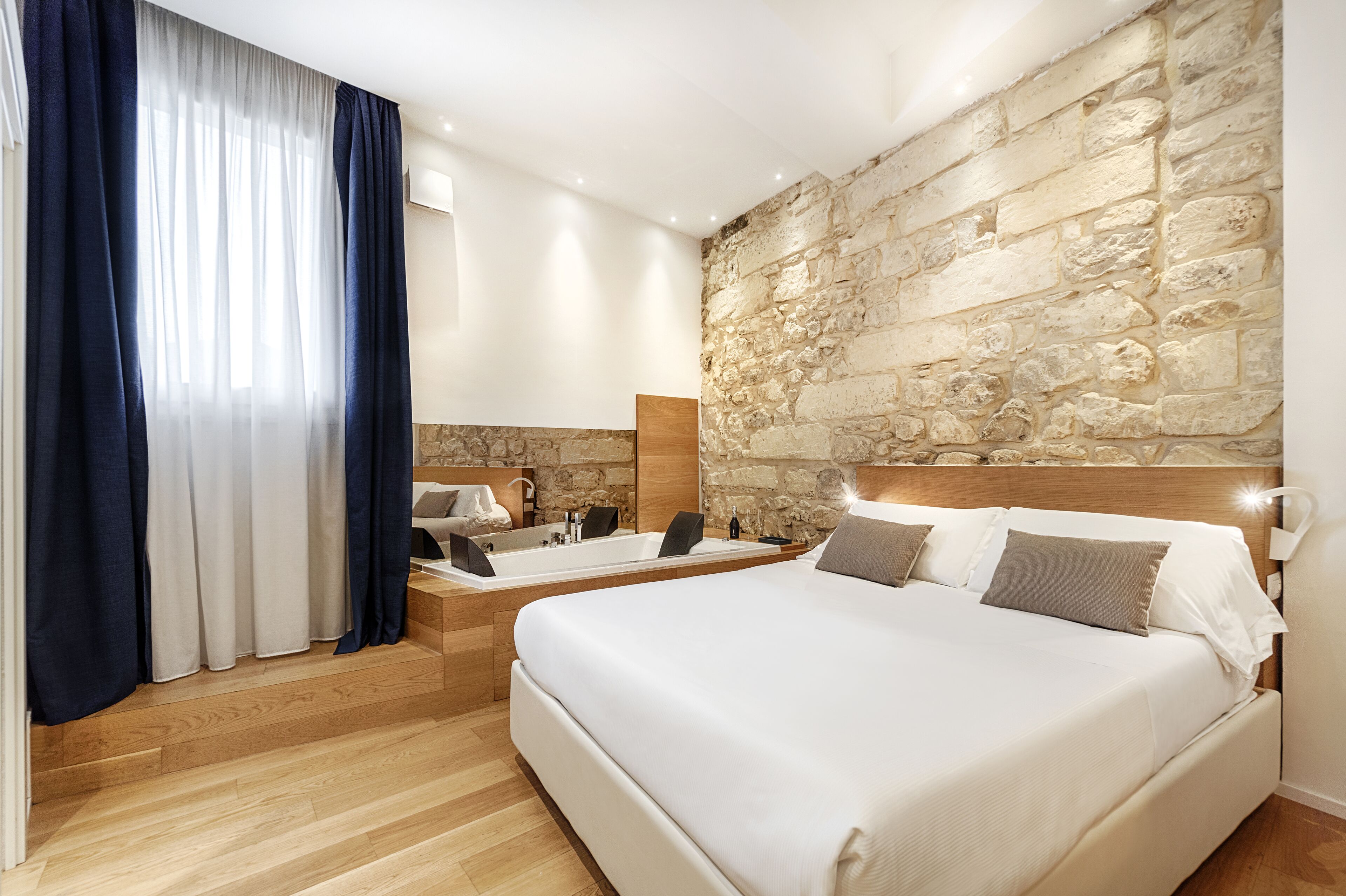 Ein modernes Schlafzimmer mit Steinwand, einem großen Bett, einer integrierten Badewanne und einem großen Fenster mit Vorhängen.