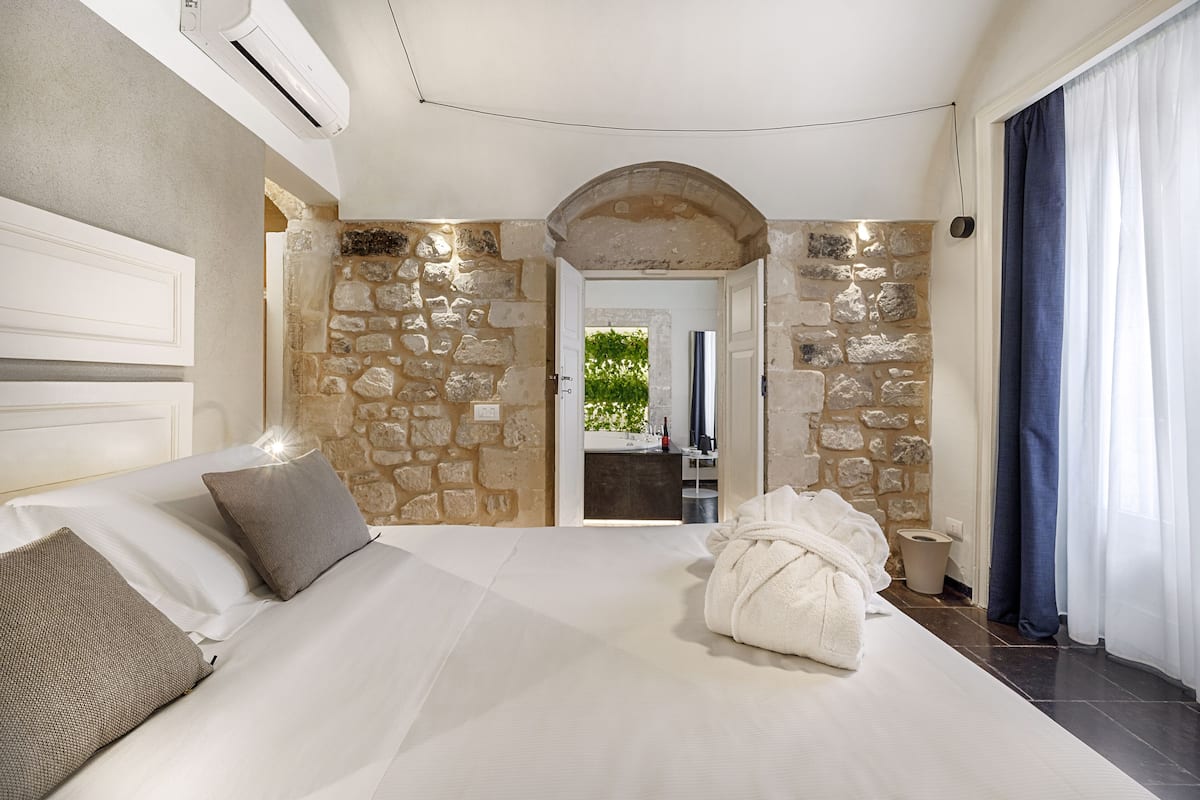 A bedroom with a stone wall, a bed with white linens, a robe on the bed, and a view of a bathroom through an archway.