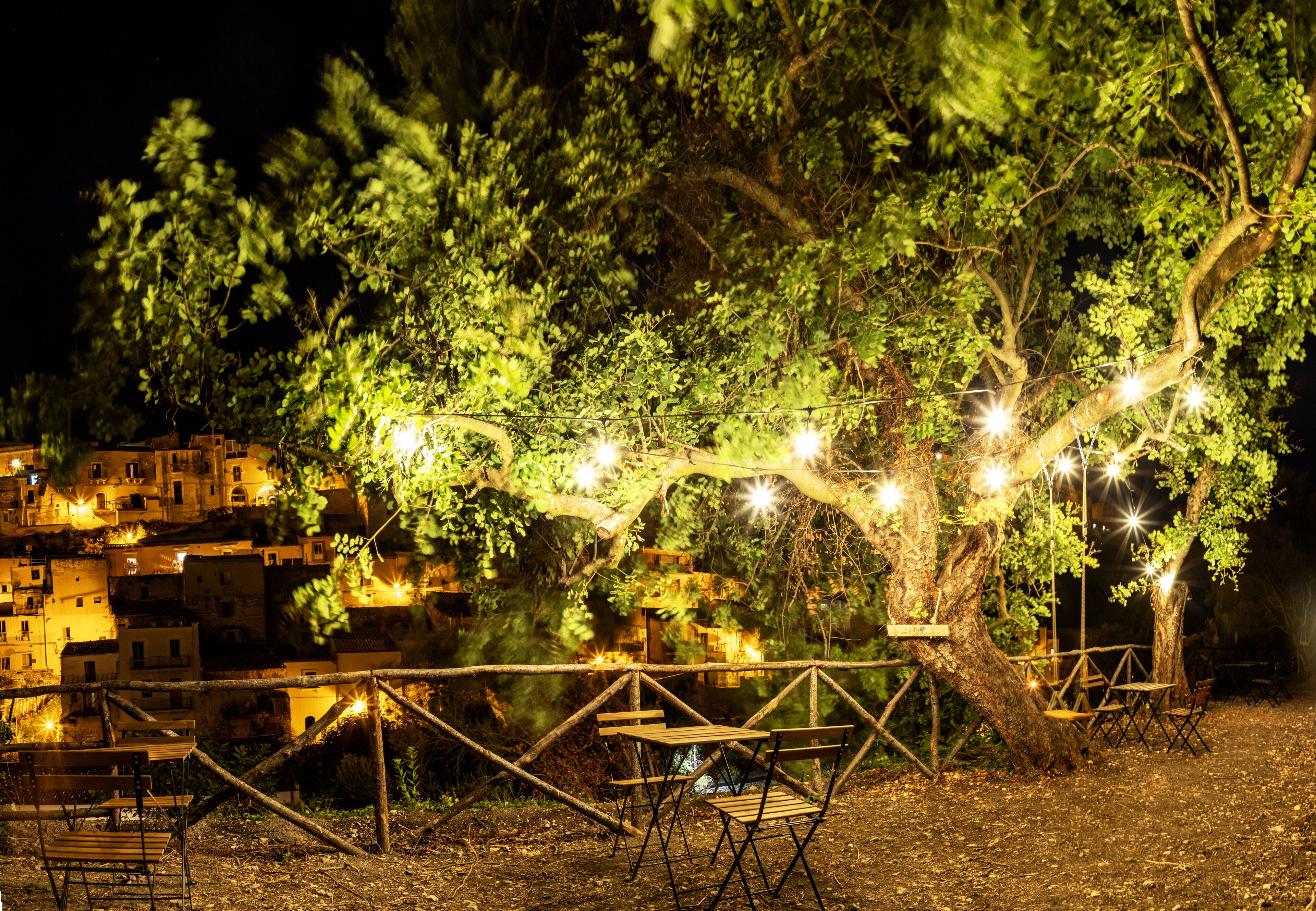 Un área de estar al aire libre con mesas y sillas bajo un árbol, rodeada por una valla, con luces colgantes y vistas a un pueblo en la ladera de noche.