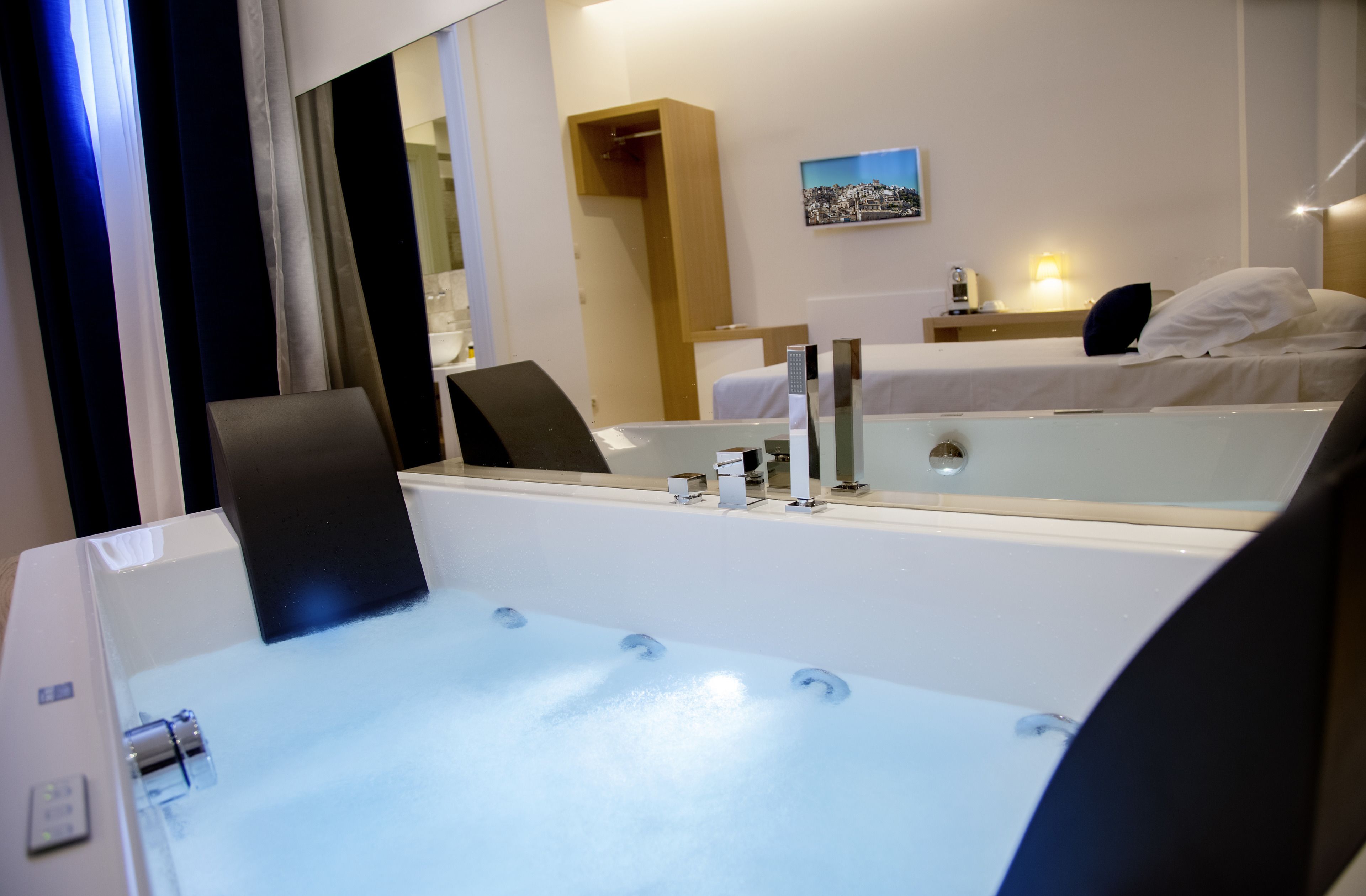 Habitación de hotel con un espejo grande, una cama, una mesita de noche con lámpara y un pequeño estante con un cuadro.