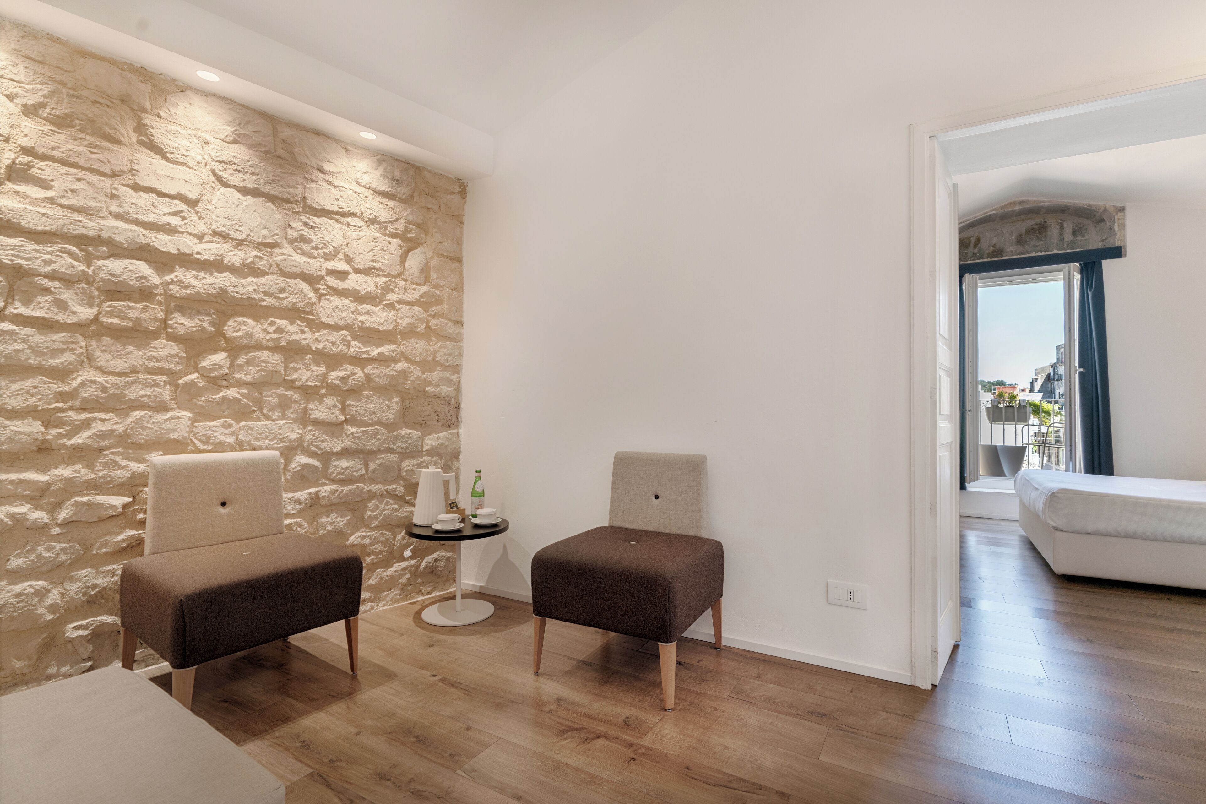 Una habitación con pared de piedra, dos sillones tapizados, una mesita redonda con una botella y un sofá blanco al fondo.