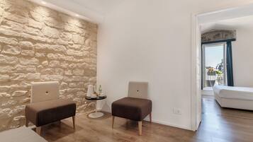 Una habitación con pared de piedra, dos sillones tapizados, una mesita redonda con una botella y un sofá blanco al fondo.