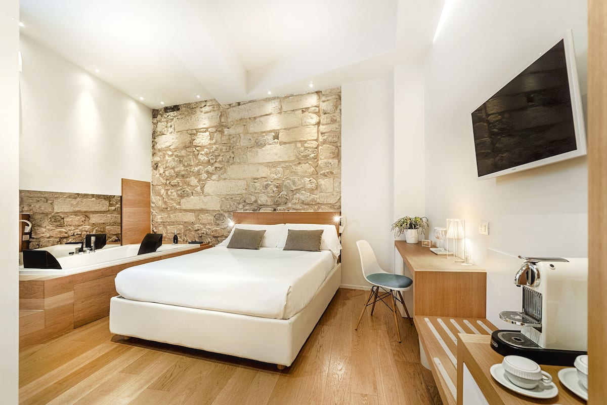 A modern bedroom with a stone wall, a large bed, a bedside table with a coffee maker, and a small round table with a lamp.