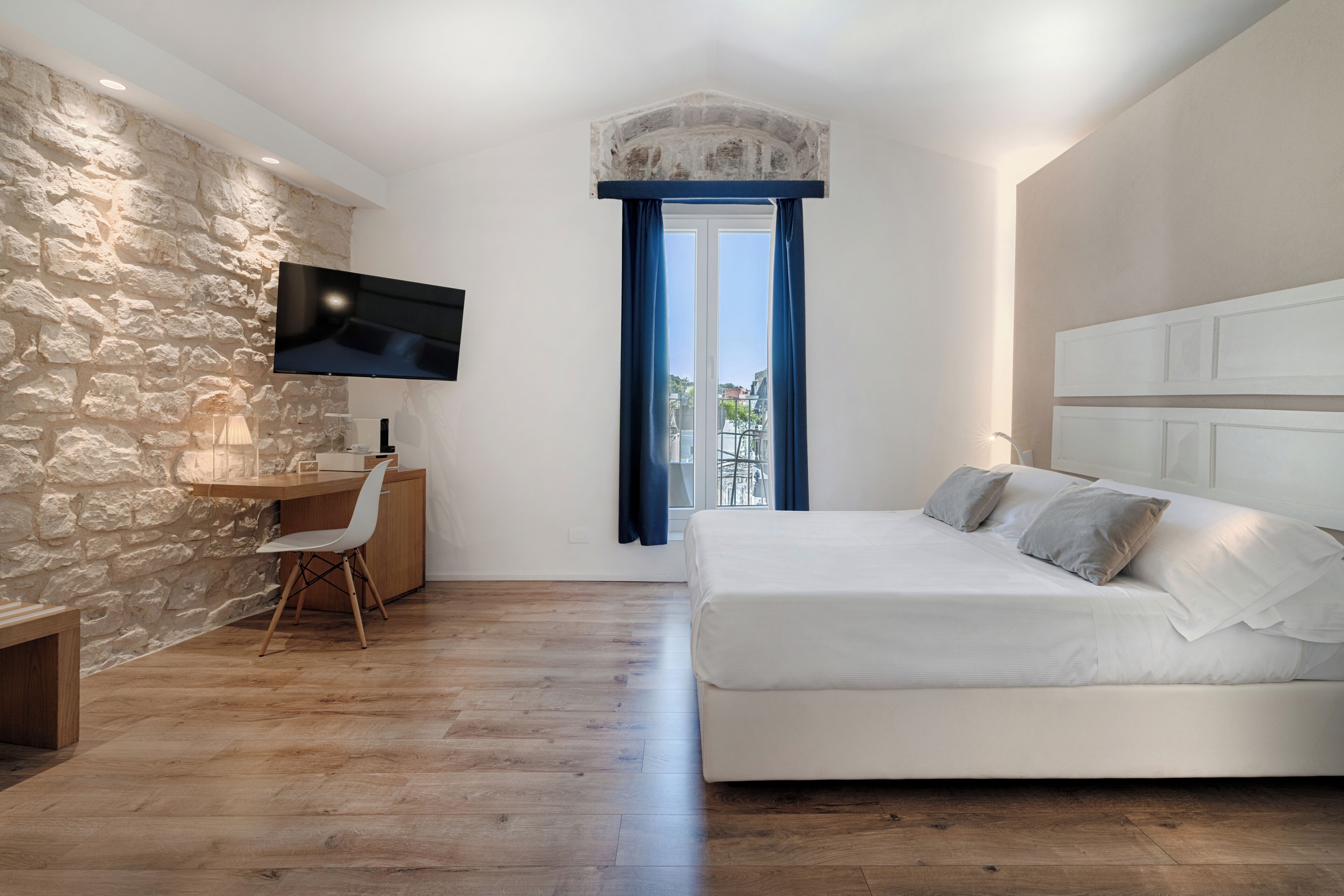 A bedroom with a stone wall, a flat-screen TV, a desk with a chair, a bed, and a view of the outdoors.