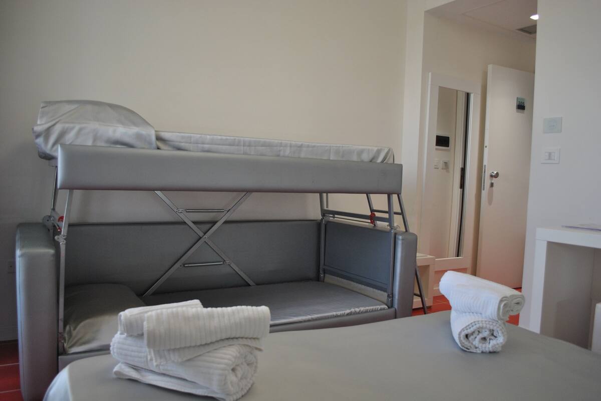 A bunk bed with a mattress and pillows, a small table with a rolled-up towel, and a door in the background.