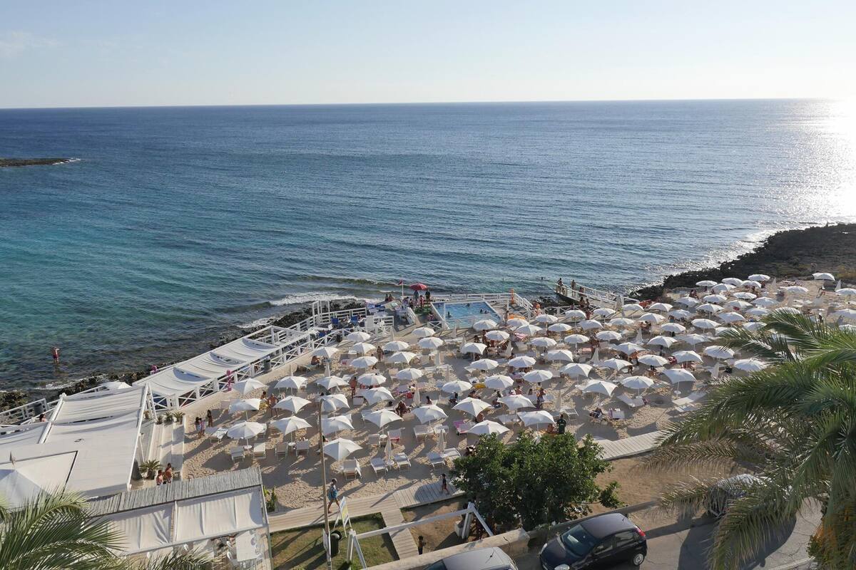 A beach with numerous white umbrellas, a pool, and a rocky shoreline.