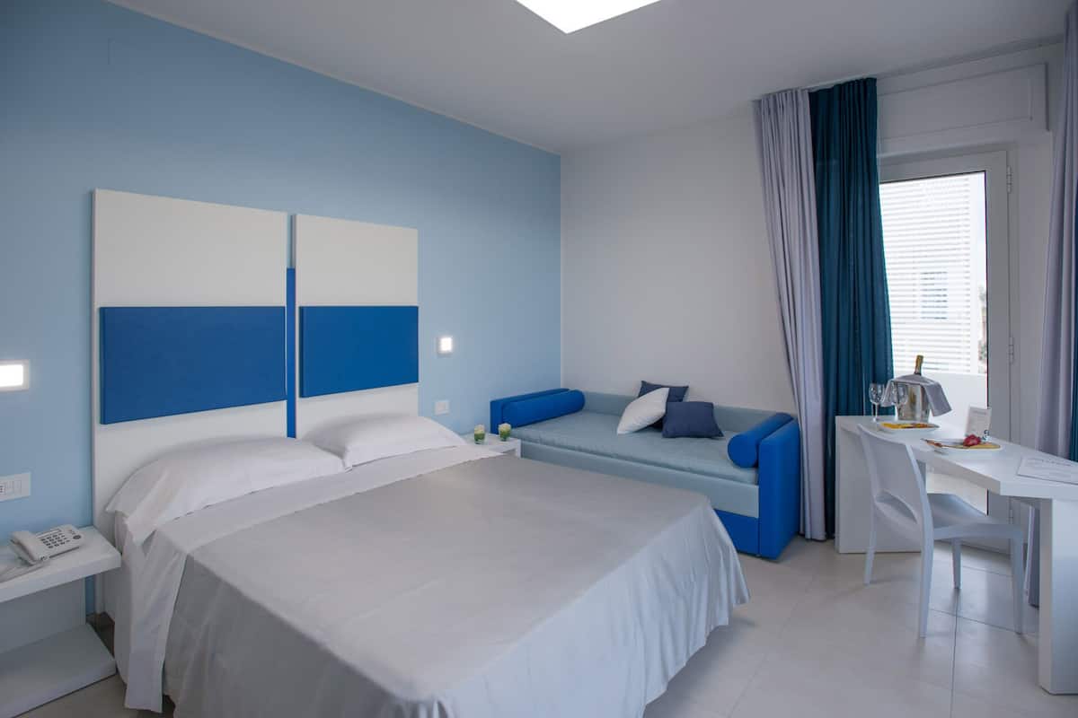 A modern hotel room with a large bed, a sofa, a desk, and a window with curtains.