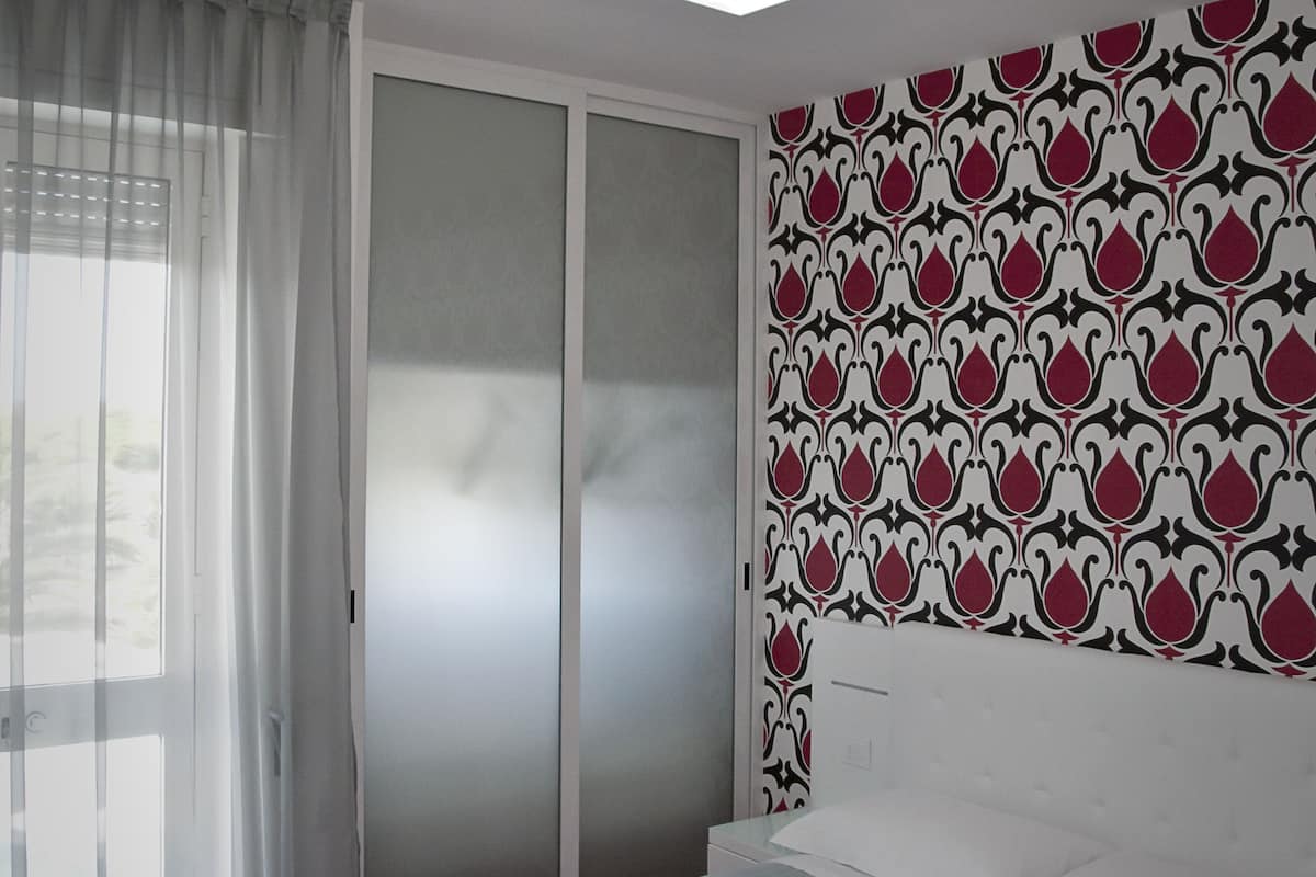 A bedroom with a bed, a wardrobe, a window with curtains, and a patterned wall.