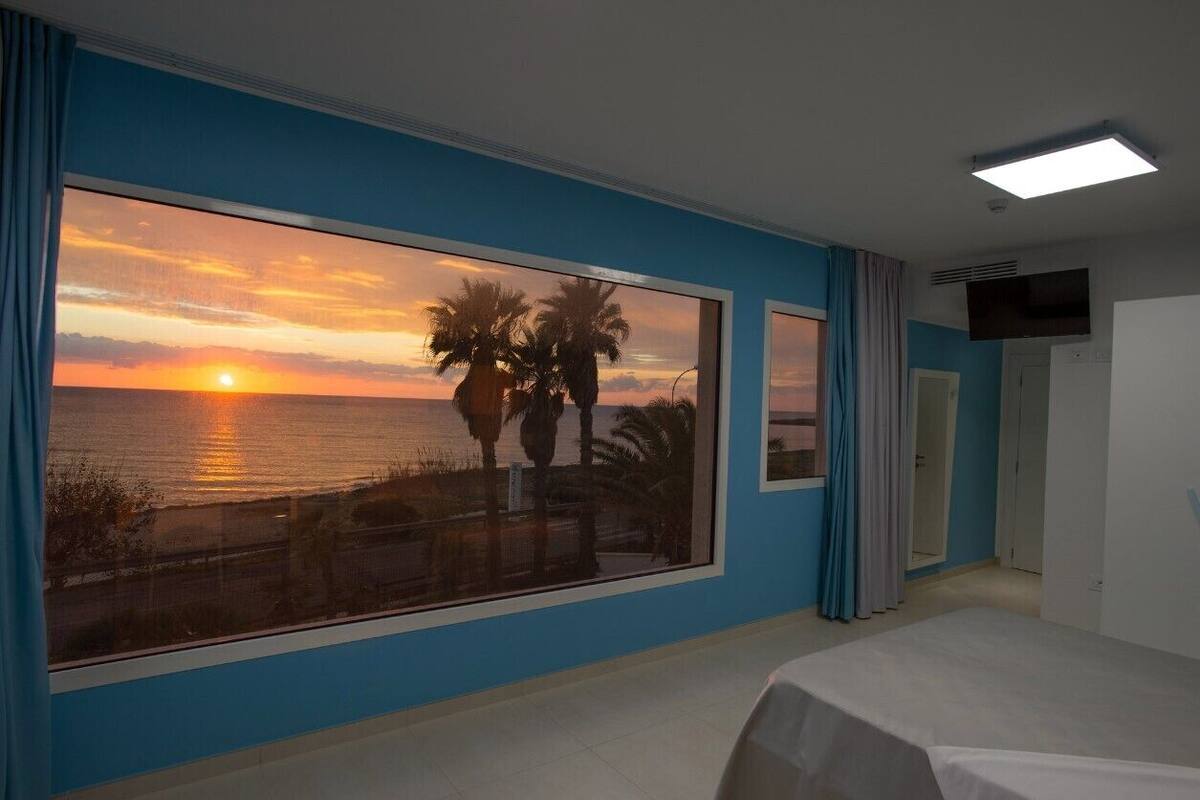 A hotel room with a large window overlooking a sunset over the ocean, a bed, and a television mounted on the wall.