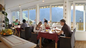 A dining area with tables and chairs, large windows with mountain views, and a variety of fruits on a table.
